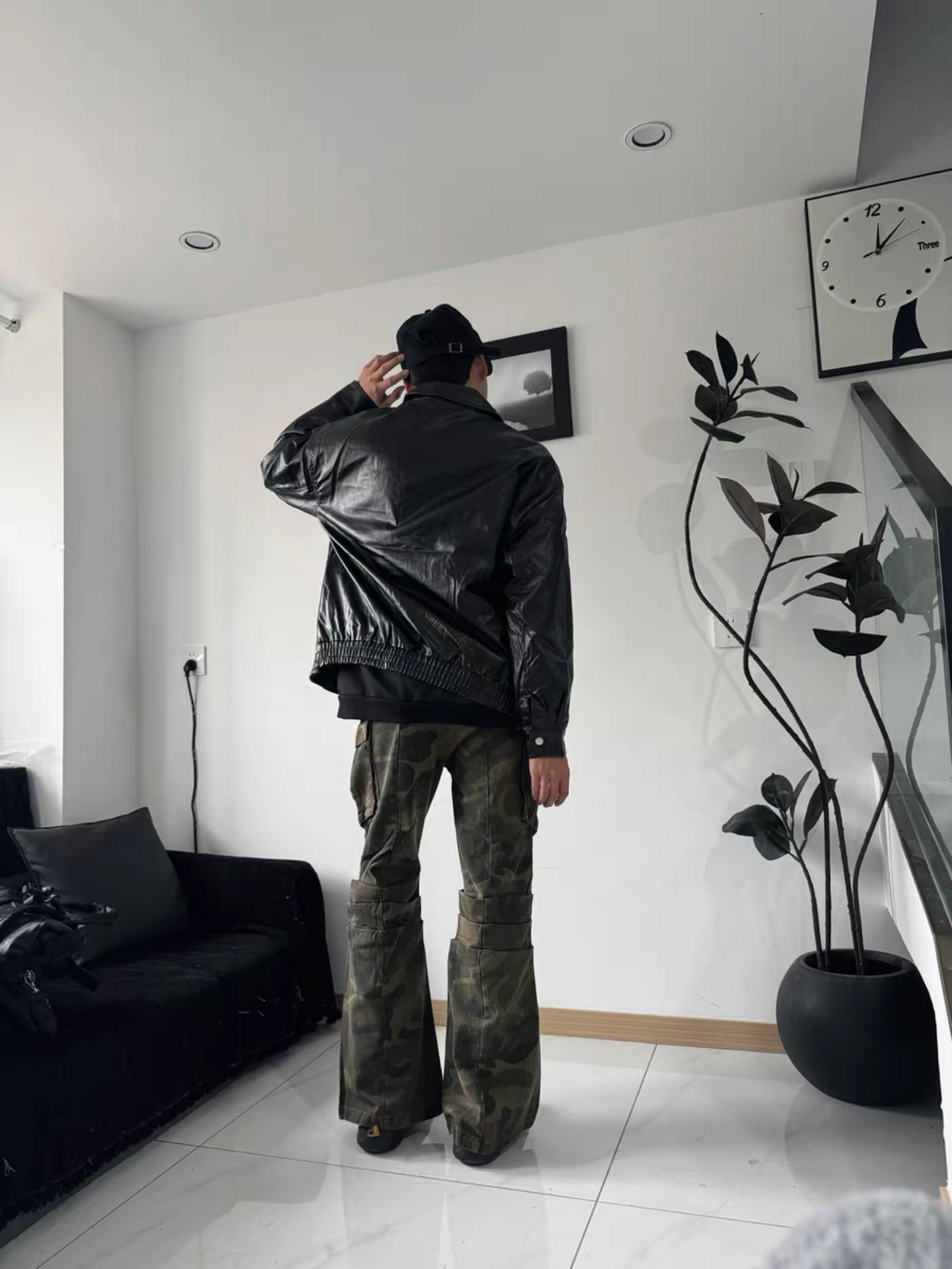 Male model showcasing back view of black PU varsity jacket with camo flared pants and cap. Indoor streetwear style with modern attitude