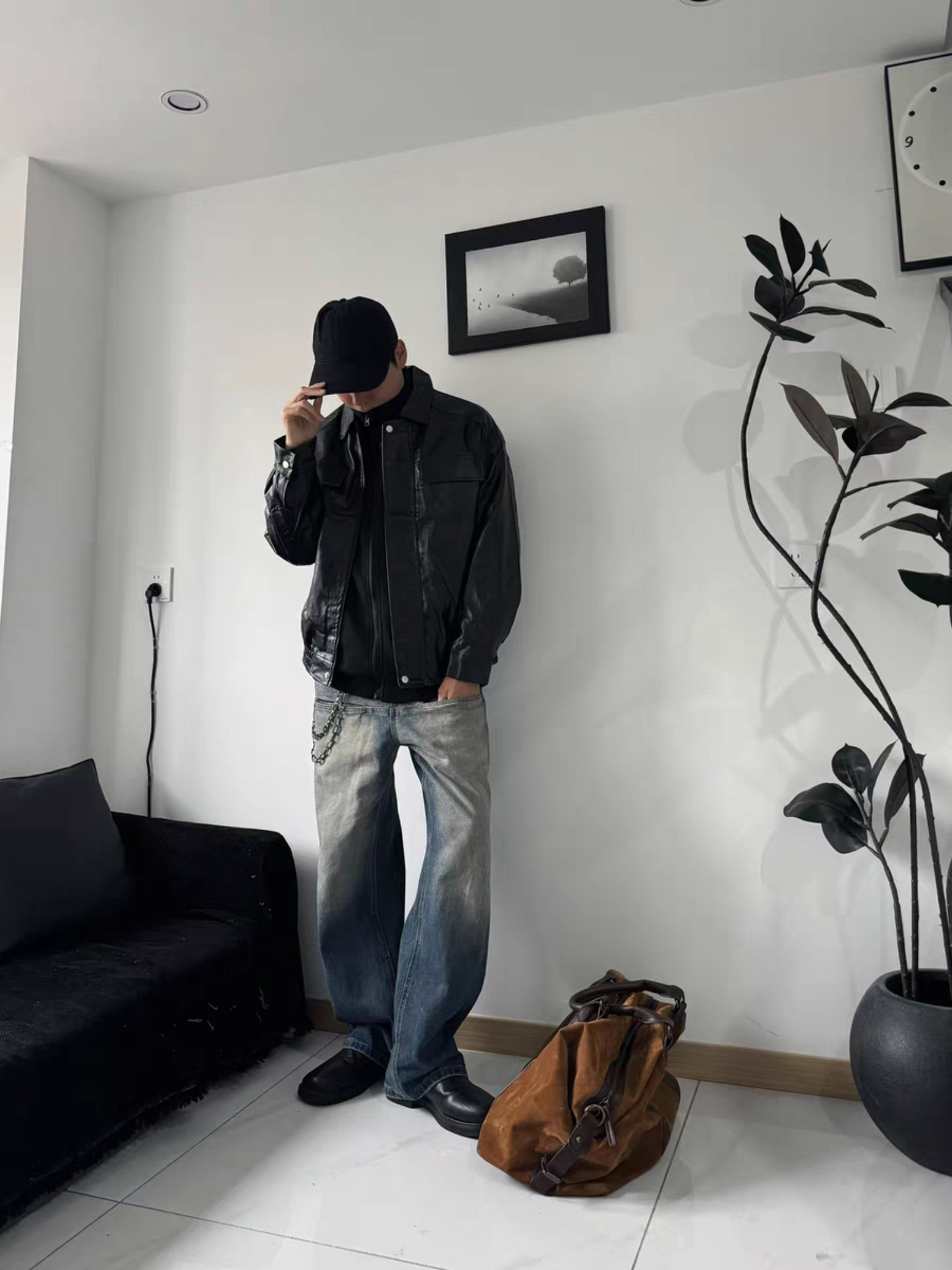 Male model wearing black PU varsity jacket with washed wide-leg jeans, cap, and leather shoes. Indoor vintage streetwear aesthetic.