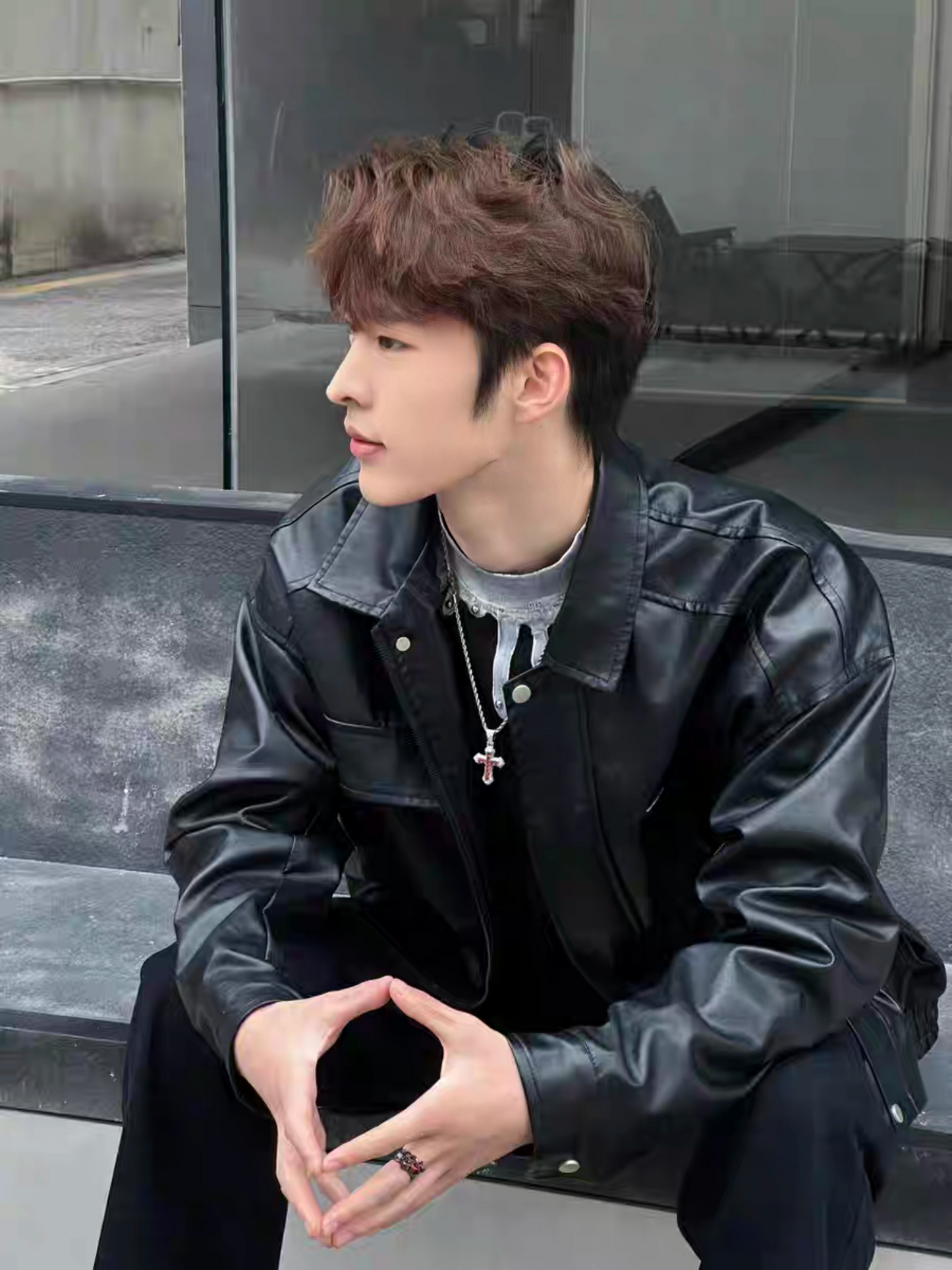 Male model in black faux leather jacket with silver cross necklace , sitting on a concrete bench outdoors showing side profile, K-style street fashion