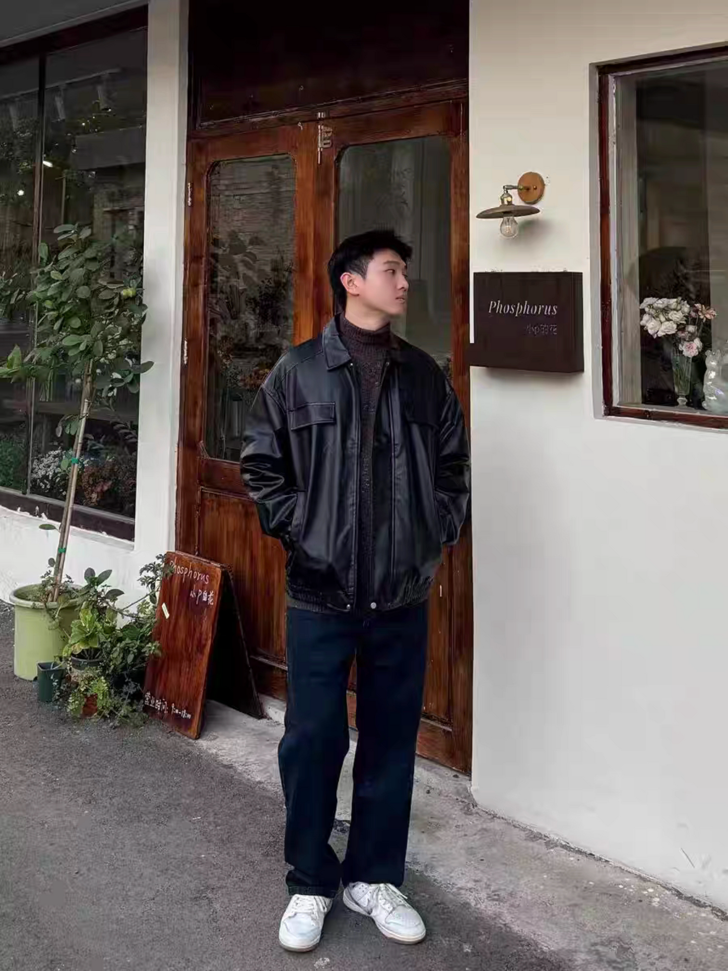 Male model wearing black PU varsity jacket standing outside a cafe, styled with dark jeans and white sneakers. Urban casual outfit inspiration.