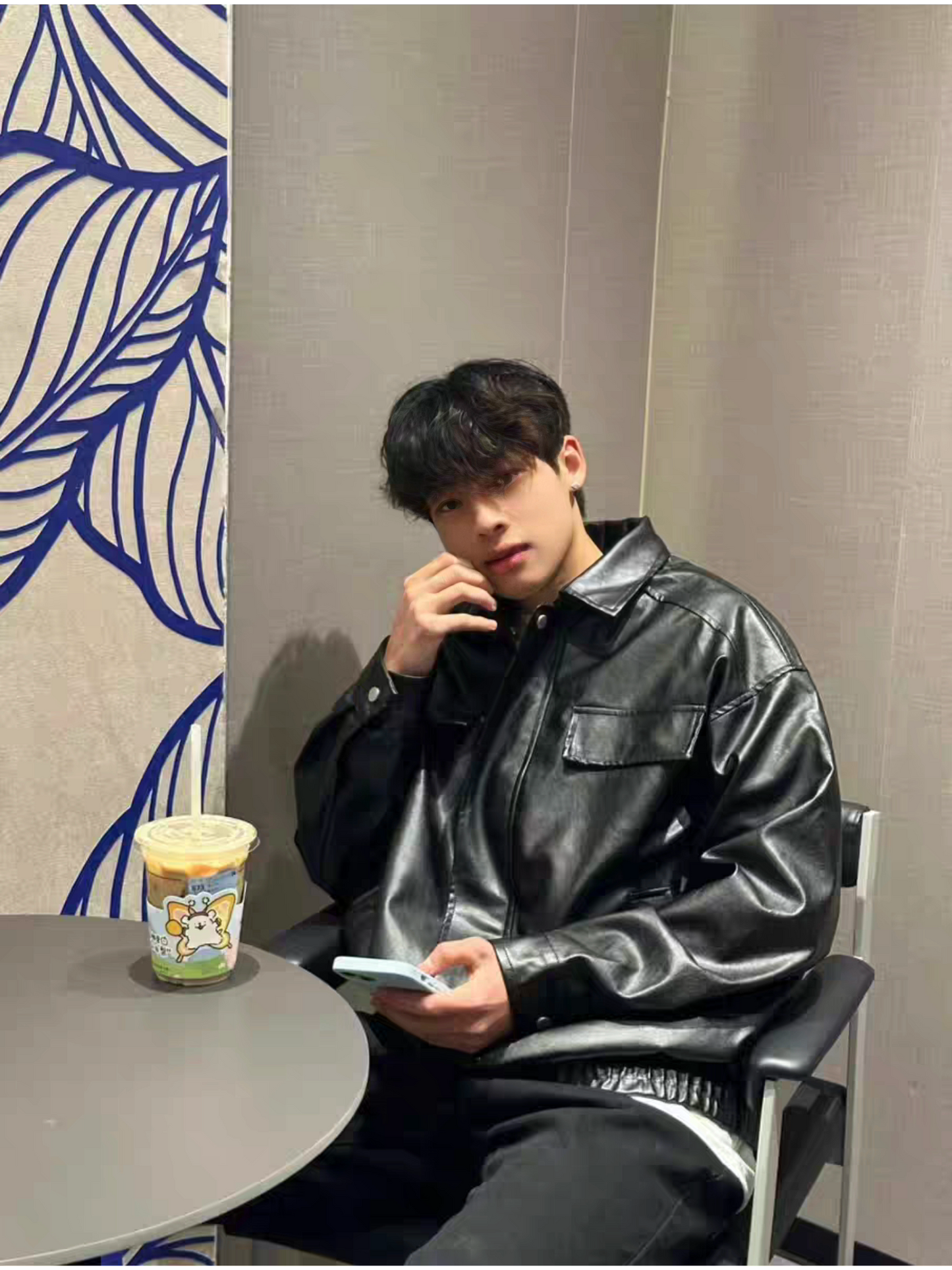 Asian male model sitting in cafe wearing STVLE black PU varsity jacket, styled casually with layered shirt and black pants, holding a phone with coffee on the table