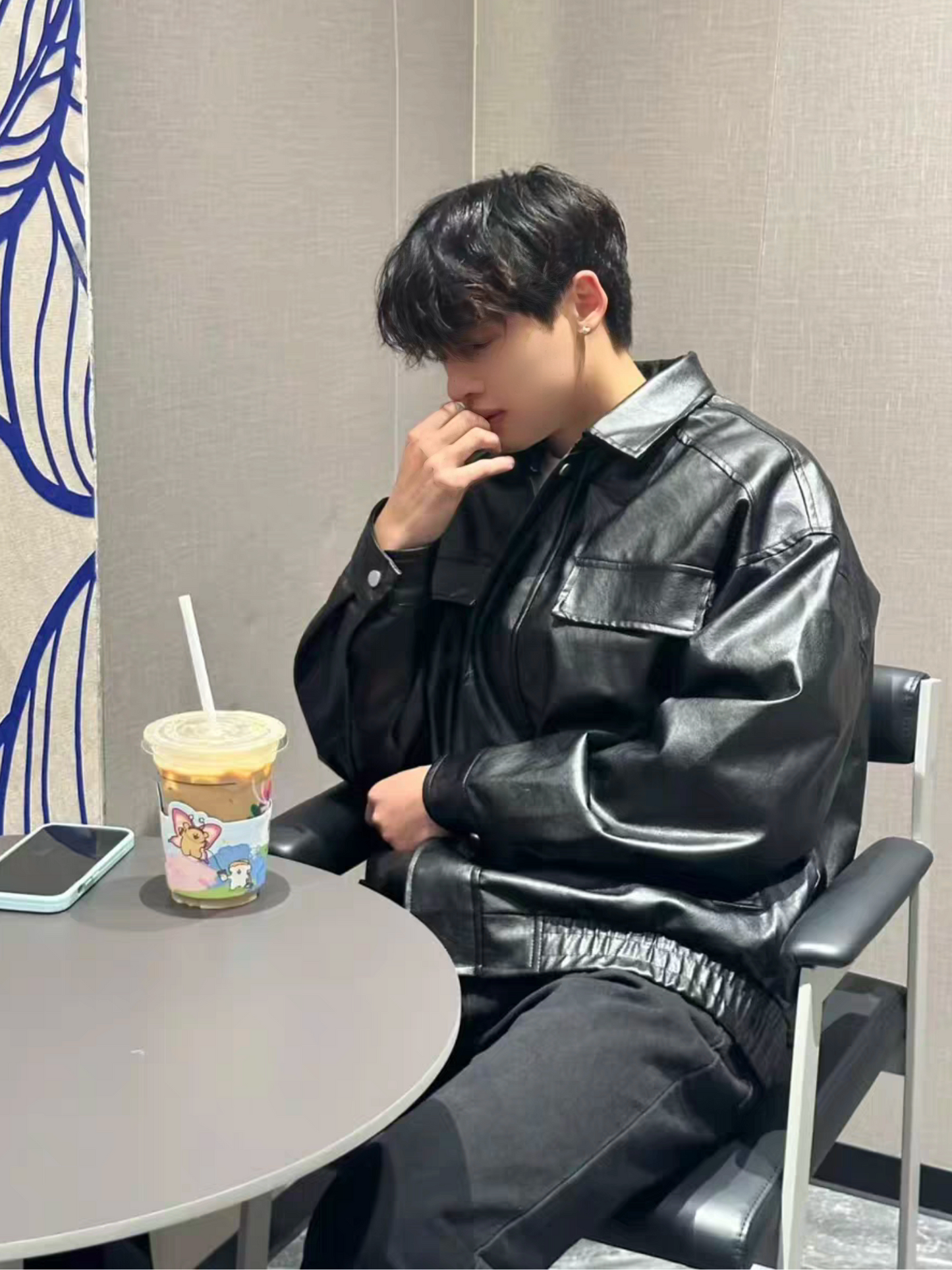 Asian male model sitting in cafe wearing STVLE black PU varsity jacket, styled with dark trousers, next to iced coffee on table. Showcasing relaxed lifestyle and urban fashion look.