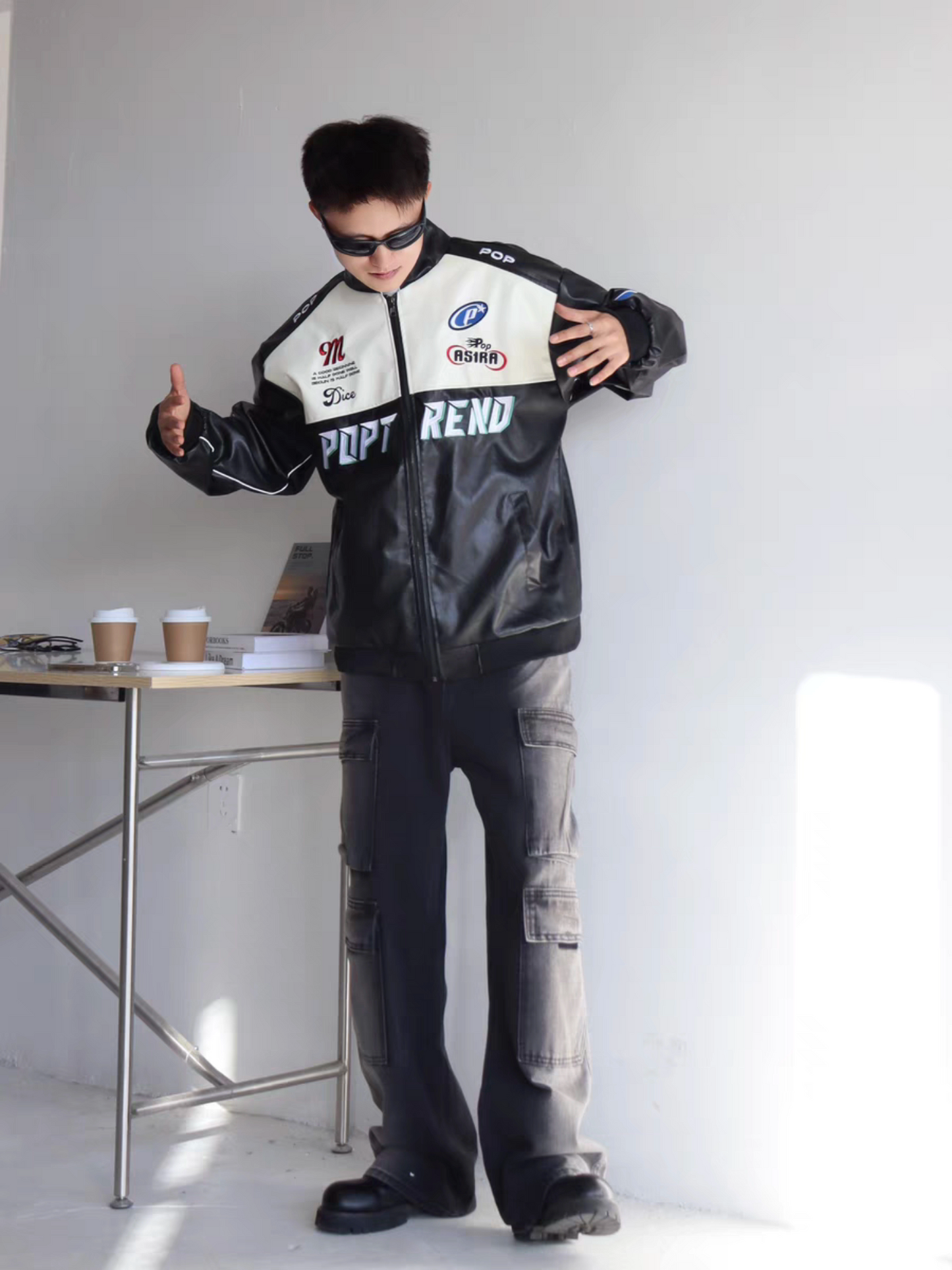 Male model in black racing-style PU leather jacket with embroidered acing patches, striking a bold pose indoors with sunglasses and cargo pants