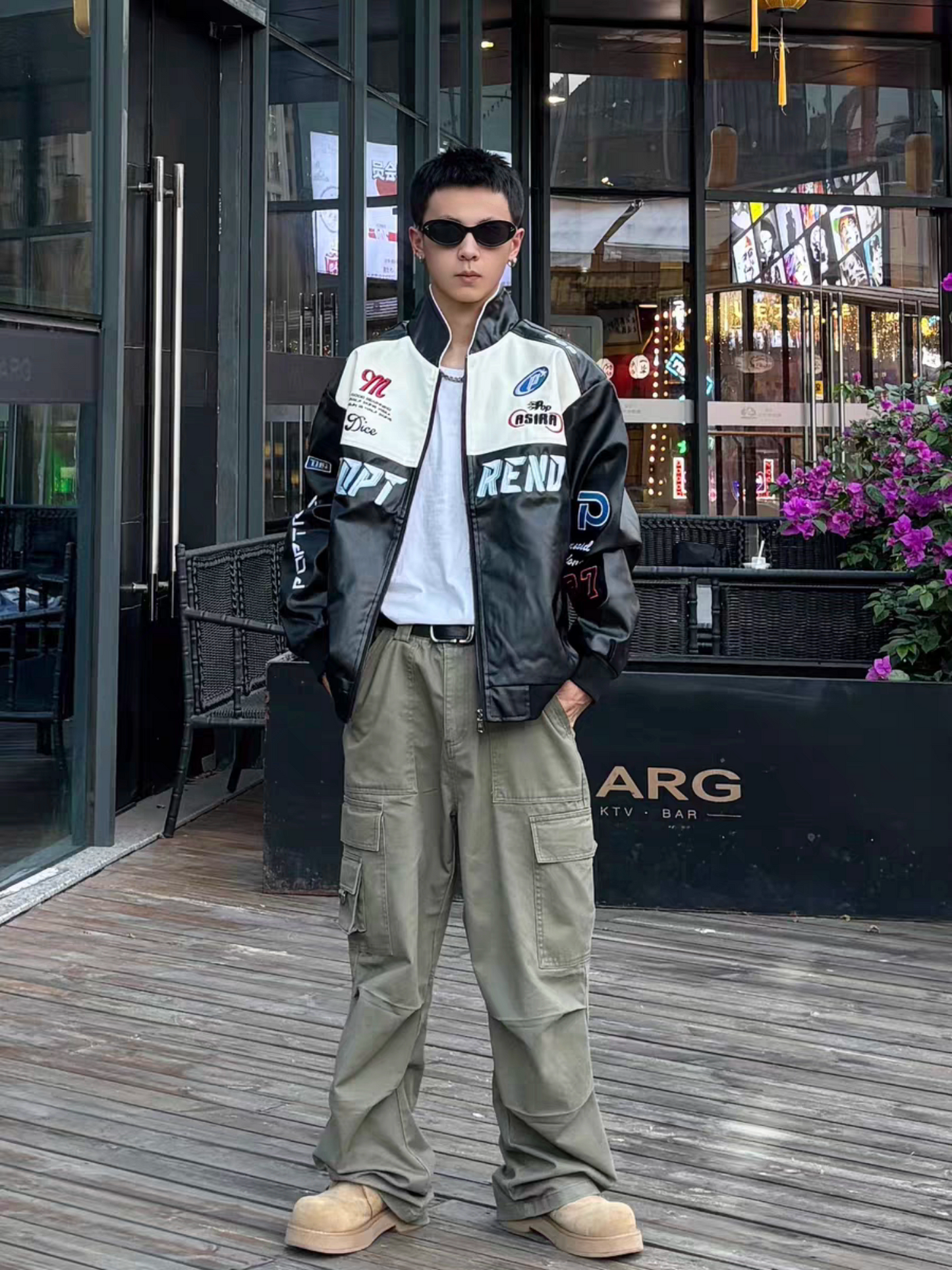 Male model standing outside a stylish bar wearing black racing PU leather jacket with sunglasses and cargo pants, showing nightlife streetwear lookj