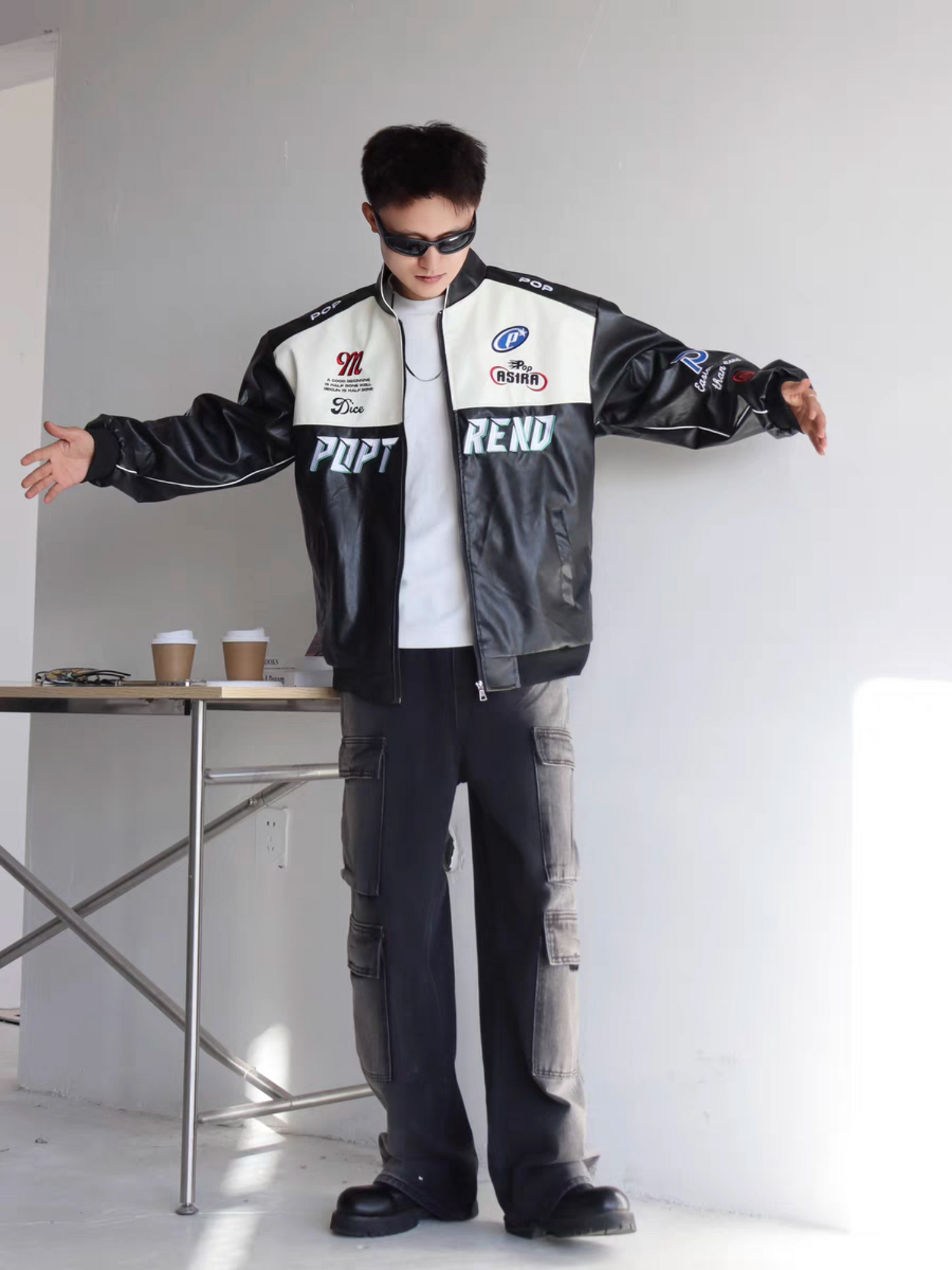 Male model wearing black racing PU jacket with arms open, showcasing relaxed fit and embroidered front details urban indoor setting