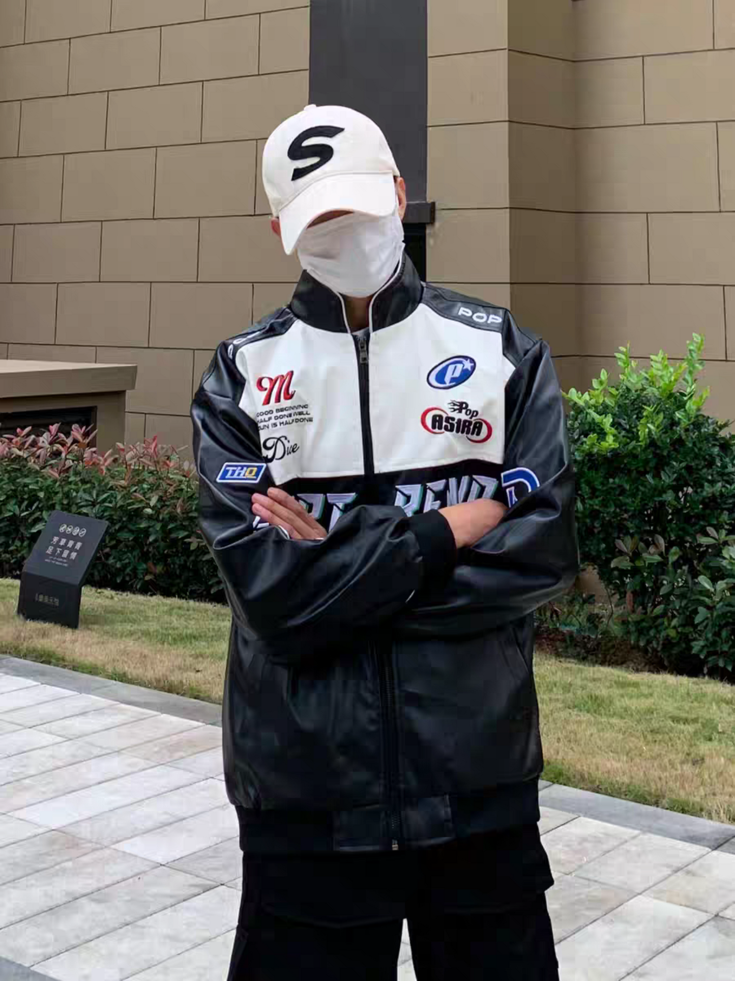 Model wearing black PU leather moto jacket with Pop Trend patches, posing outdoors with arms crossed and cap