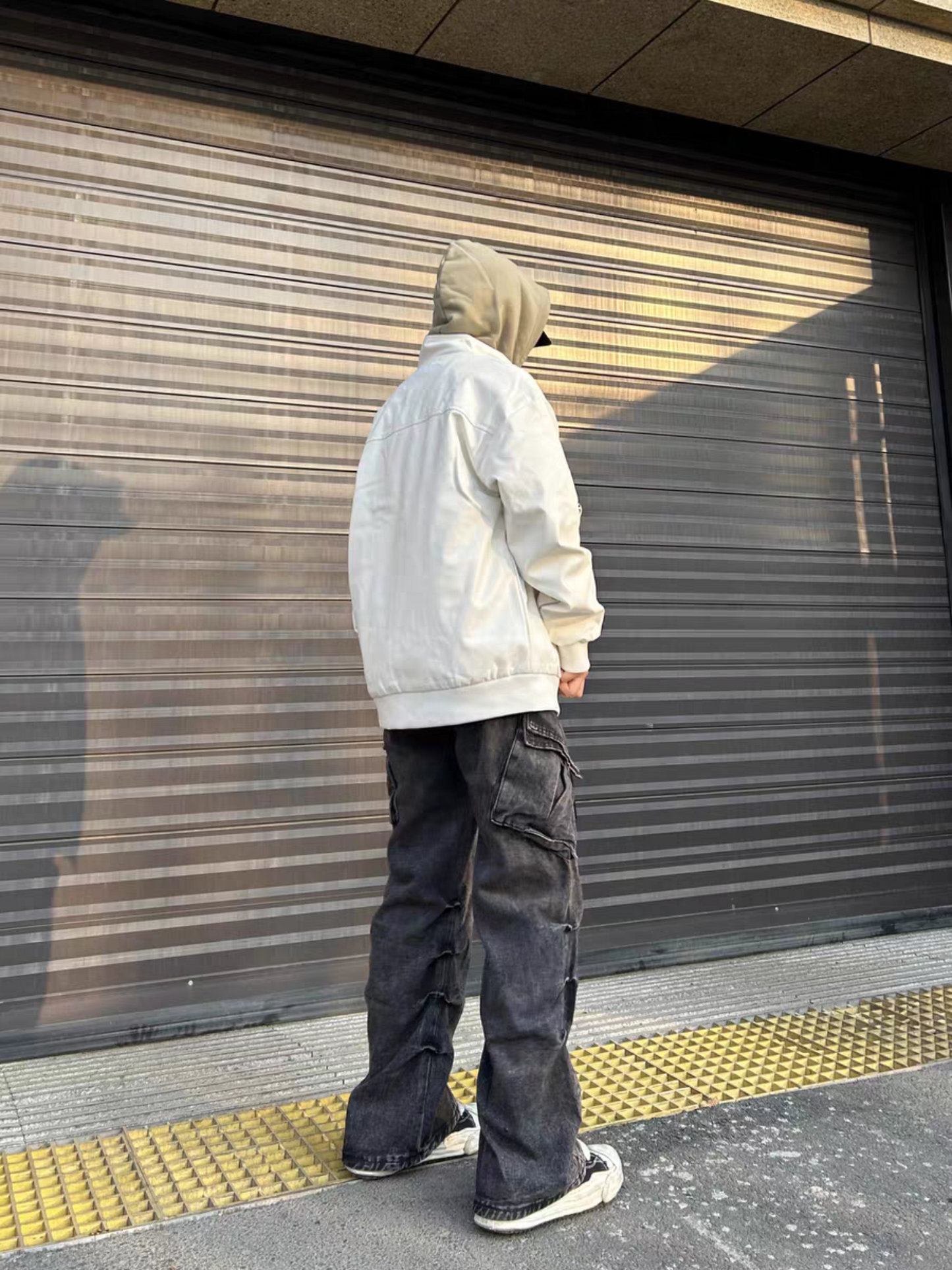 Back view of a model wearing a white racing jacket with a hoodie layered underneath, styled with loose black cargo pants and sneakers on urban street
