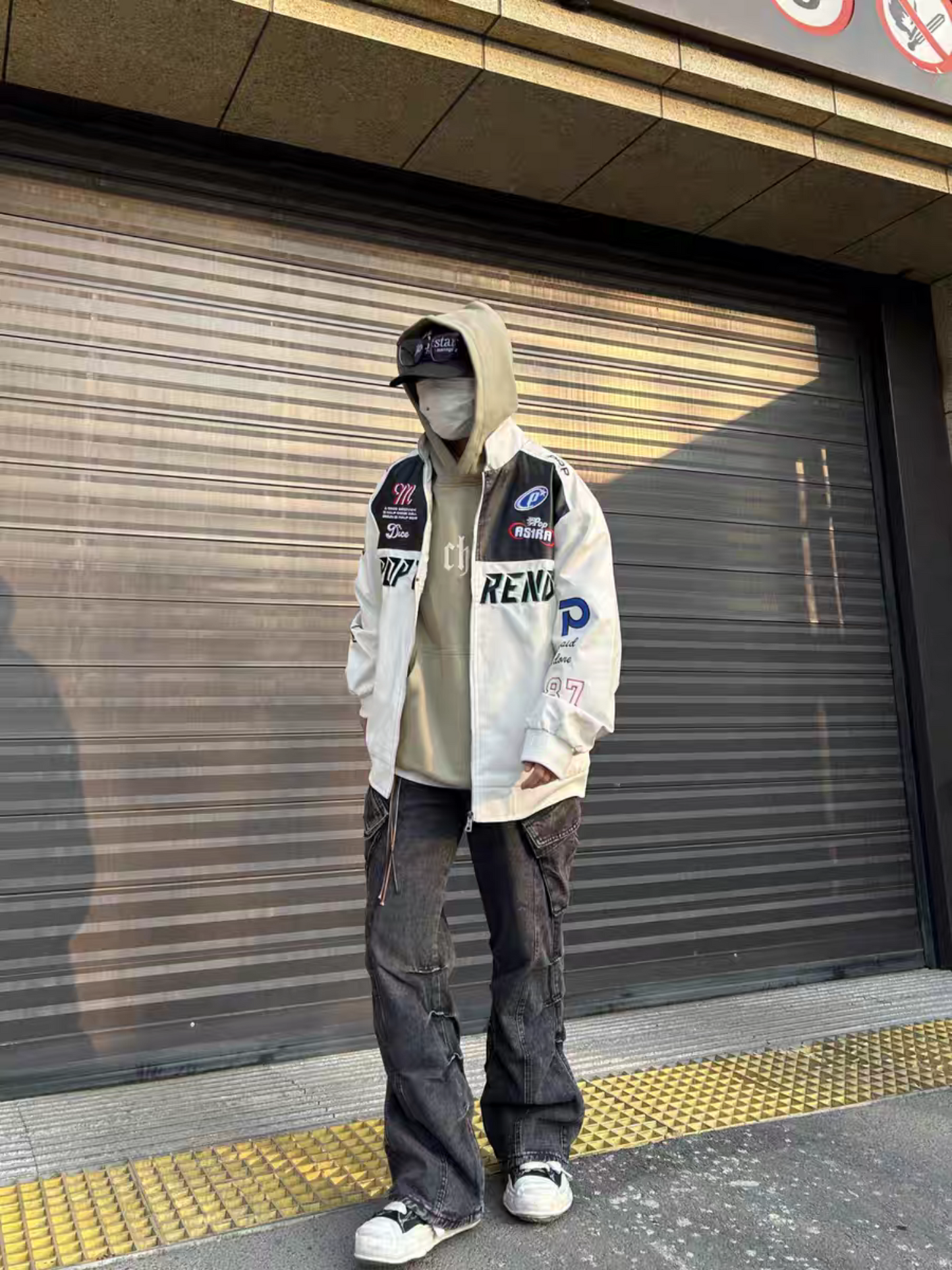 Male model wearing white racing PU leather jacket in a streetwear outfit , paired with hoodie and cargo pants, standing in front of a shutter gate