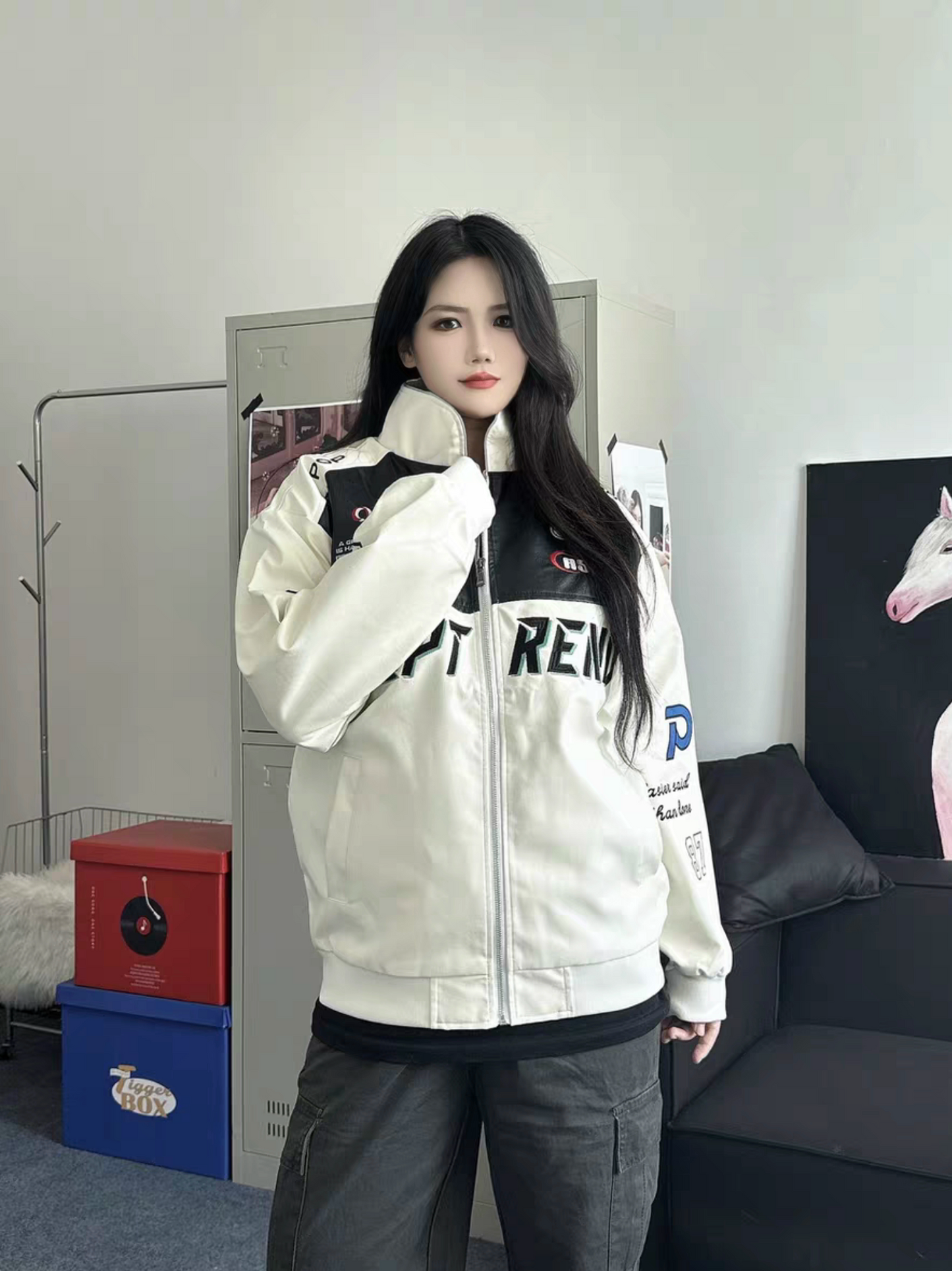Female model wearing a white racing style moto jacket with embroidered logos, standing indoors in a minimalistic room with lockers and modern decor