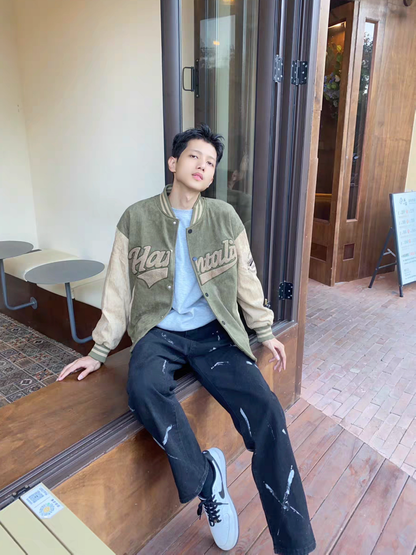 Male model sitting casually in green and brown Hantaliv varsity jacket, styled with black paint-splattered jeans and sneakers in an outdoor cafe setting