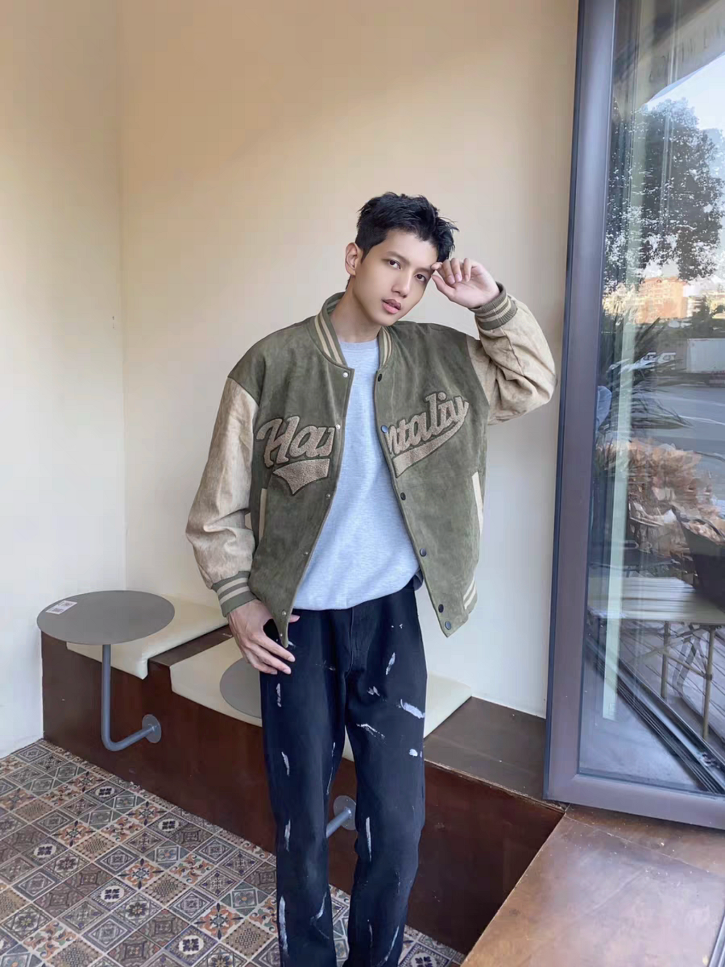 Male model posing in green and brown Hantaliv jacket next to a window, paired with distressed black jeans and gray tee in a lifestyle setting
