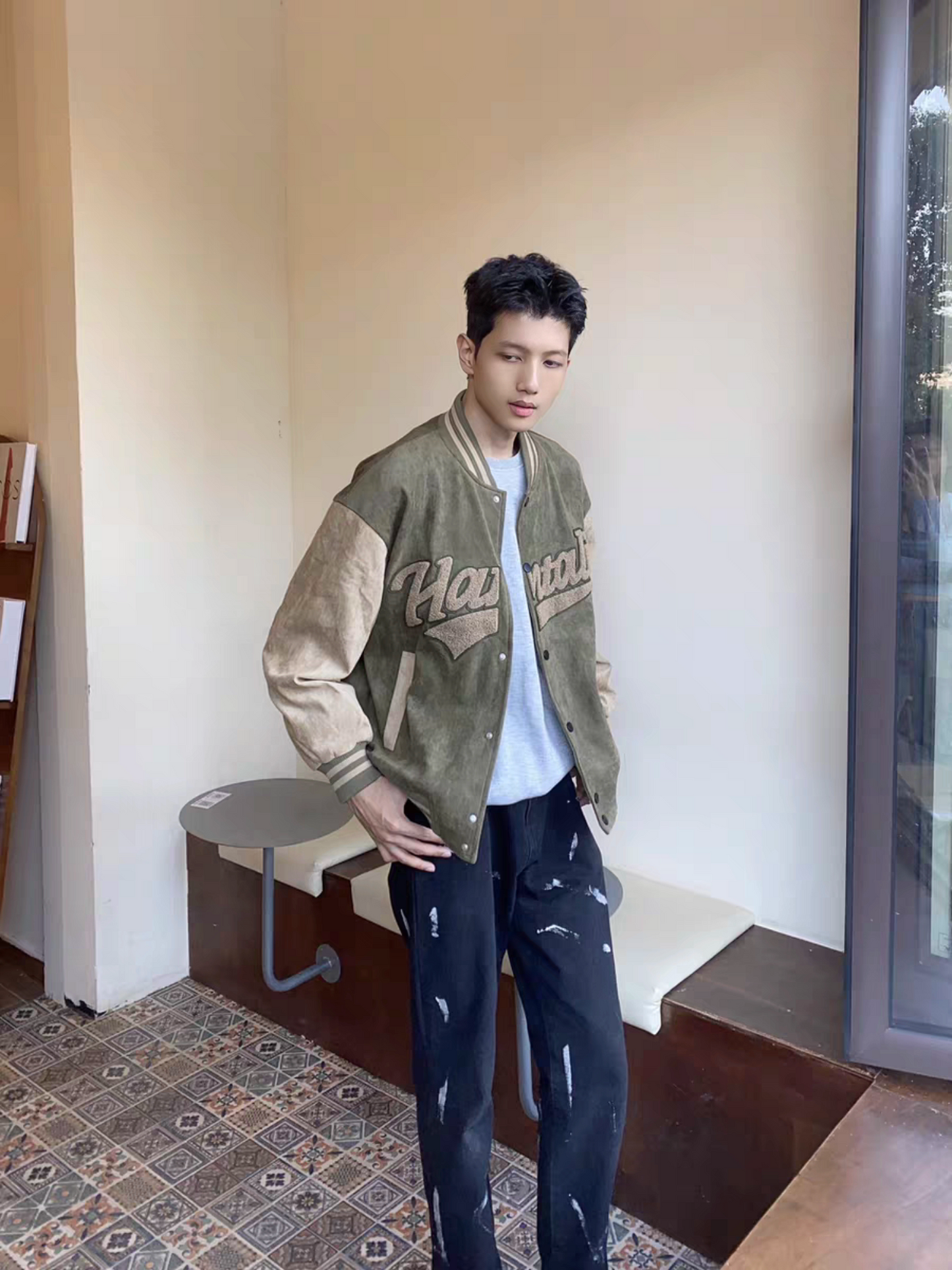 Male model wearing green and brown Hantaliv varsity jacket standing indoors near a window, styled with distressed black jeans and grey T-shirt