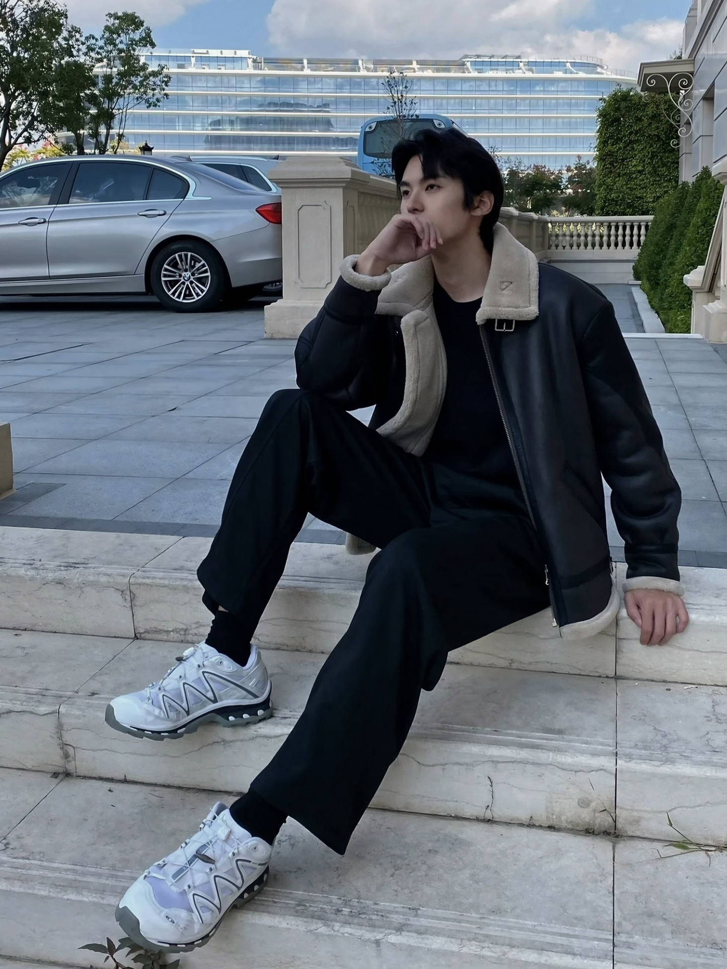 Man wearing black faux shearling aviator jacket with beige lining, sitting casually on marble steps outdoors