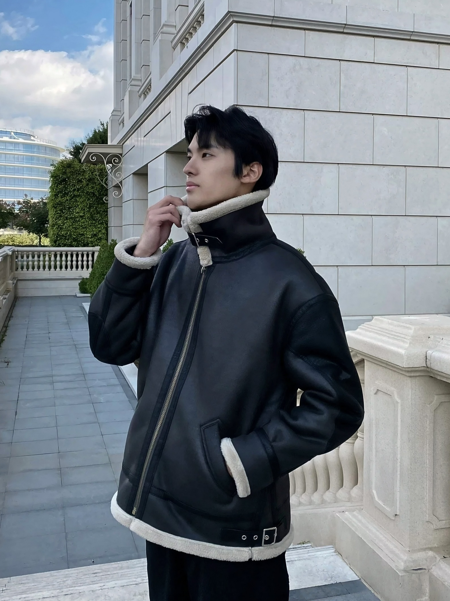 Man wearing black faux shearling aviator jacket with buckle strap collar, standing in front of European-style building