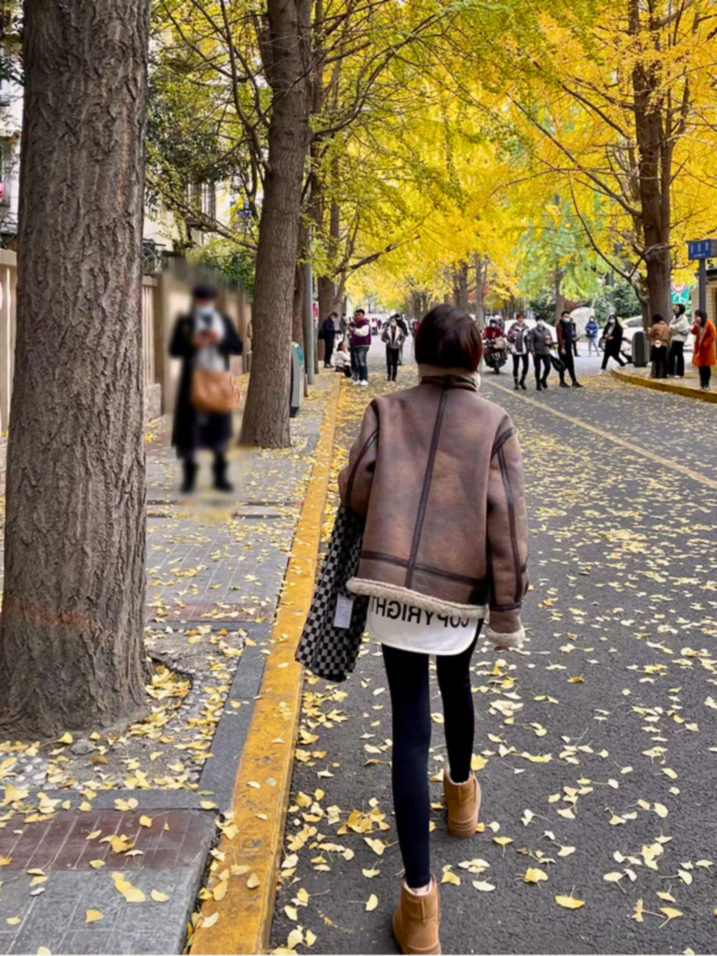 Woman in brown faux shearling aviator jacket walking down autumn street lined with golden ginkgo trees