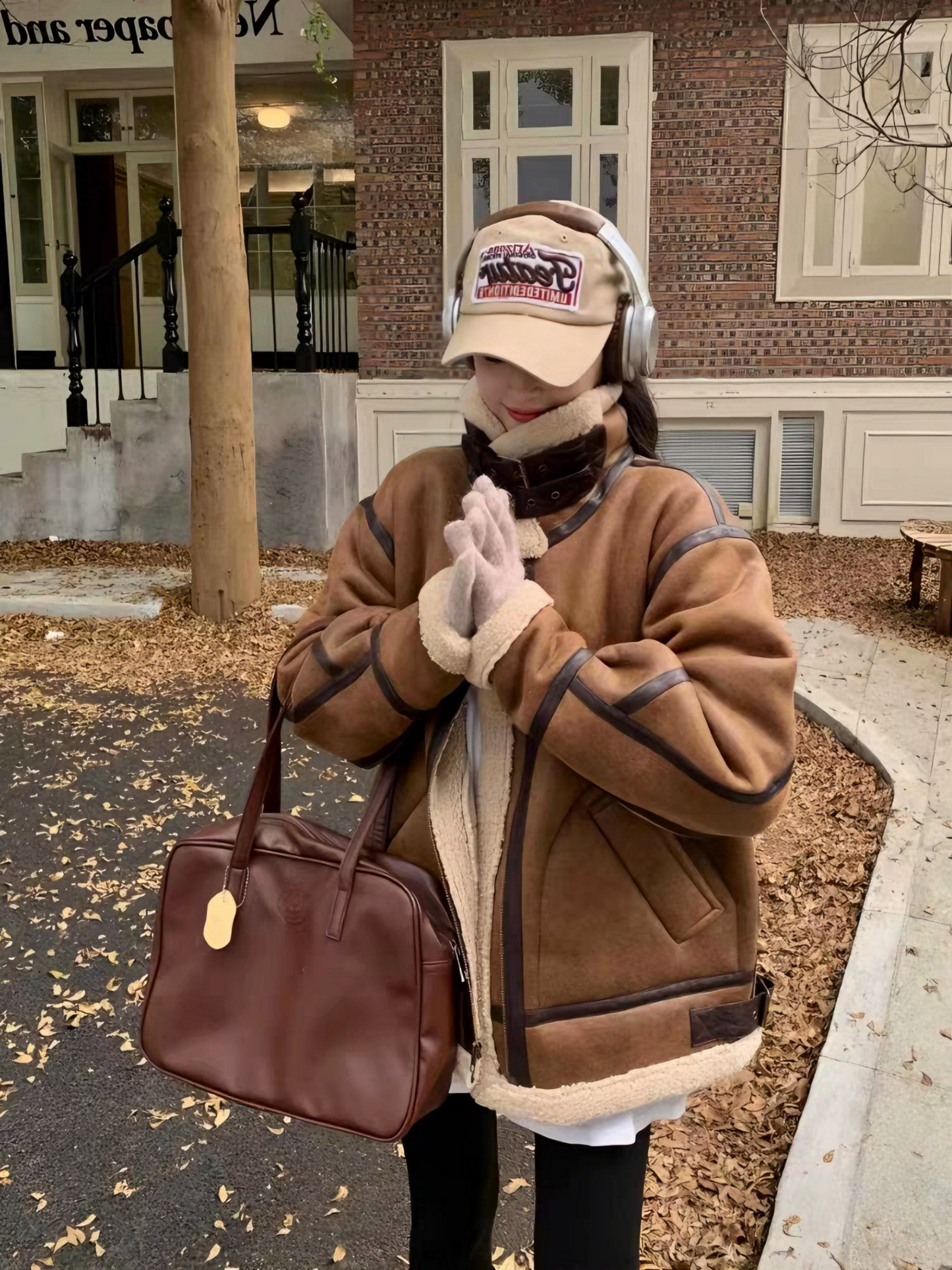 Woman in brown faux shearling aviator jacket with headphones and cap, holding hands together in autumn street setting