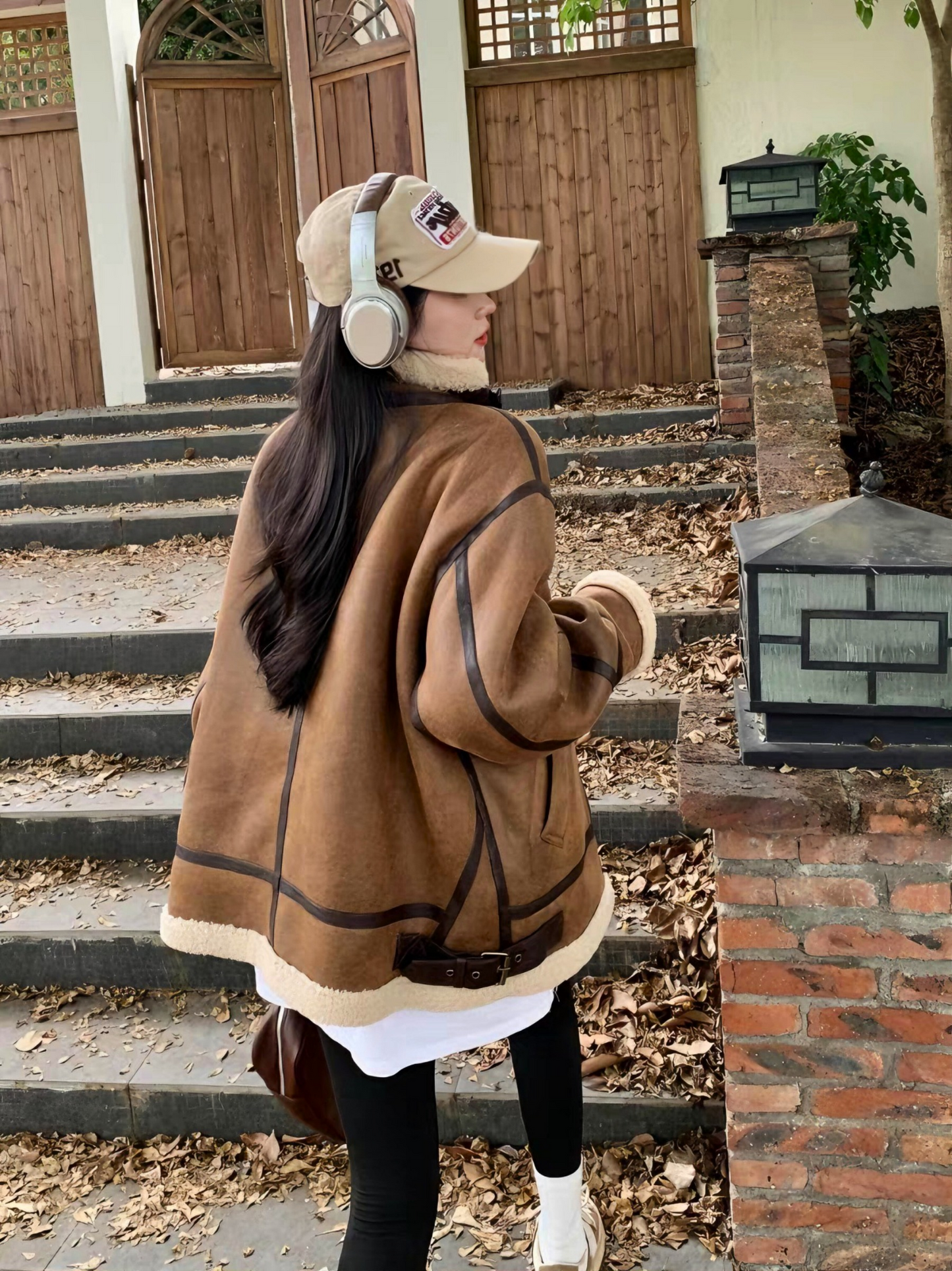 Back view of woman in brown faux shearling aviator jacket with cap and headphones, walking up outdoor stairs in autumn leaves