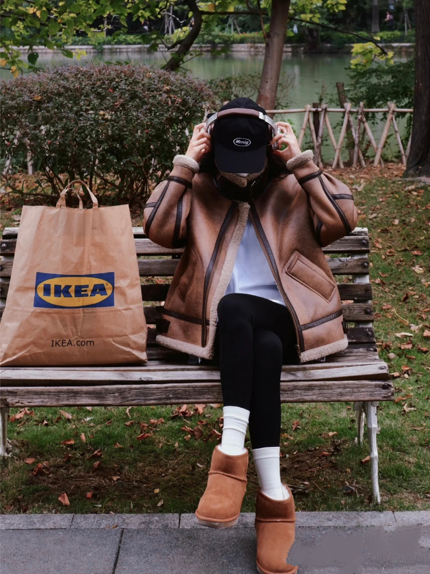 Woman wearing brown faux shearling aviator jacket adjusting headphones while sitting on park bench with IKEA bag