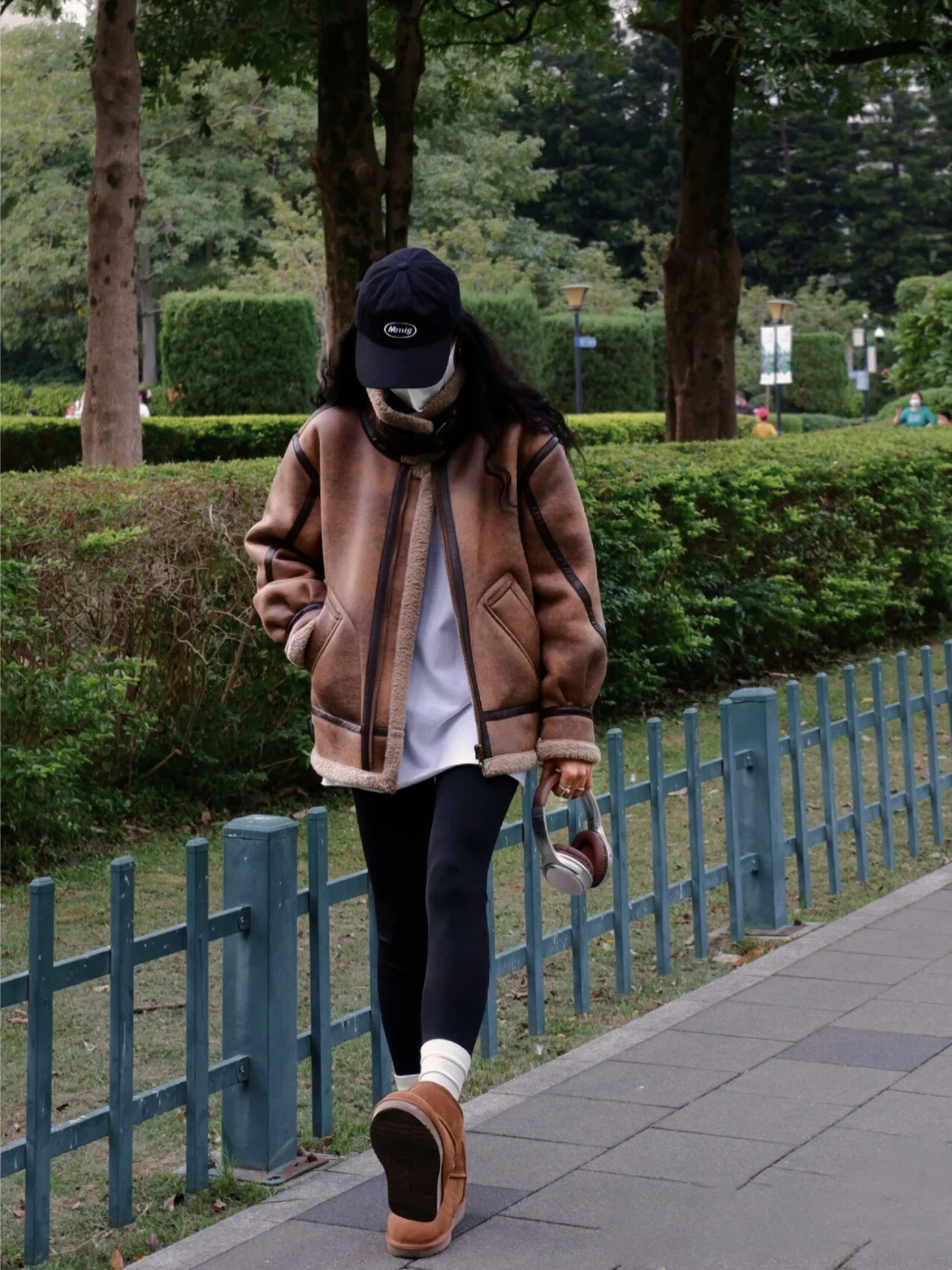 Woman in brown faux shearling aviator jacket walking in park with casual winter outfit and cap
