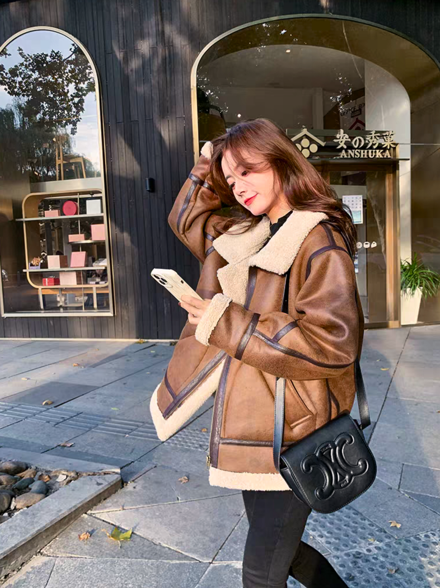 Woman wearing brown faux shearling aviator jacket, checking phone in sunny street style look