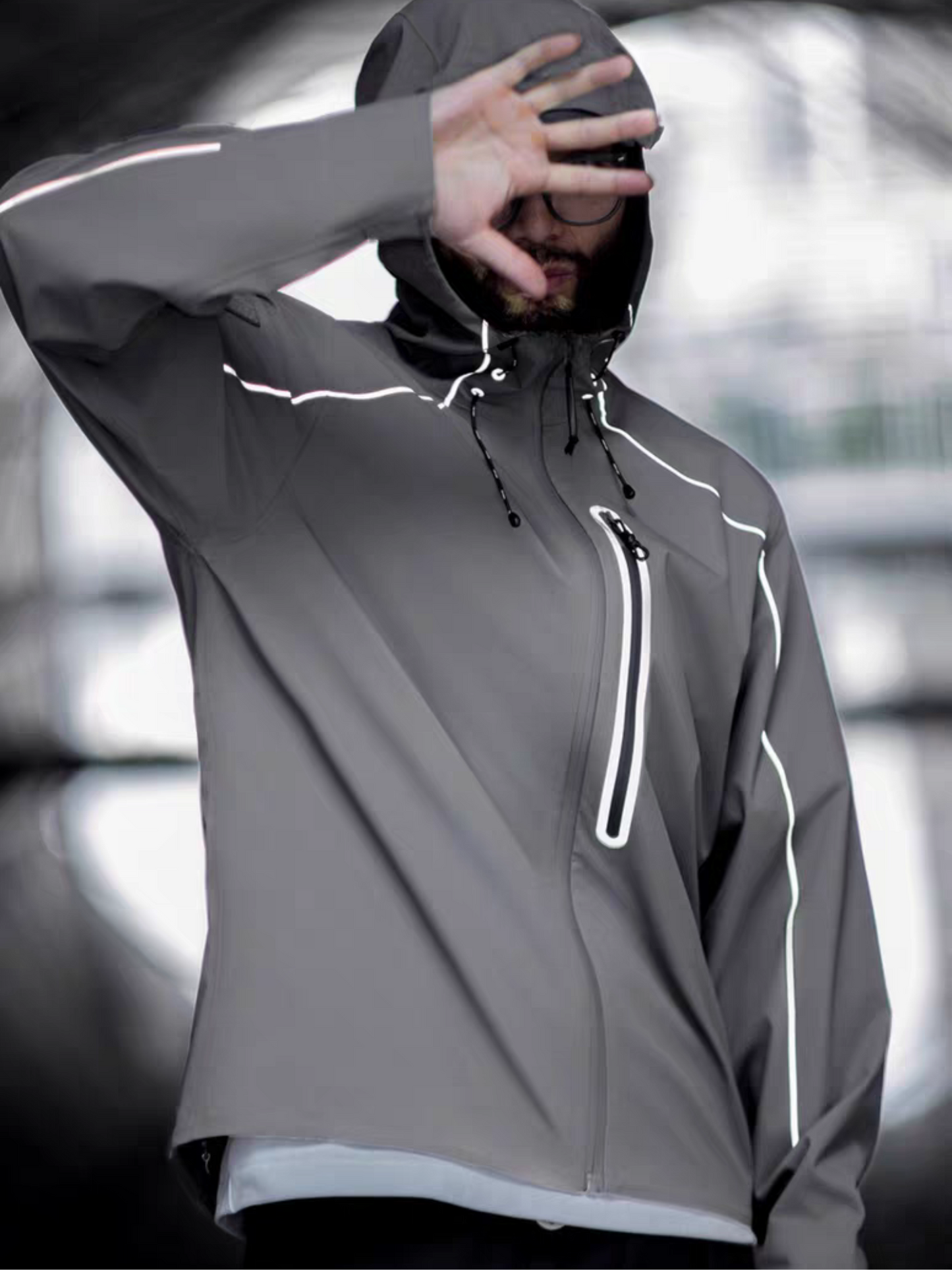 Model wearing STVLE lightsaber reflective techwear jacket, hood on, hand covering face - -shot under urban overpass with tactical posture and minimalist design details