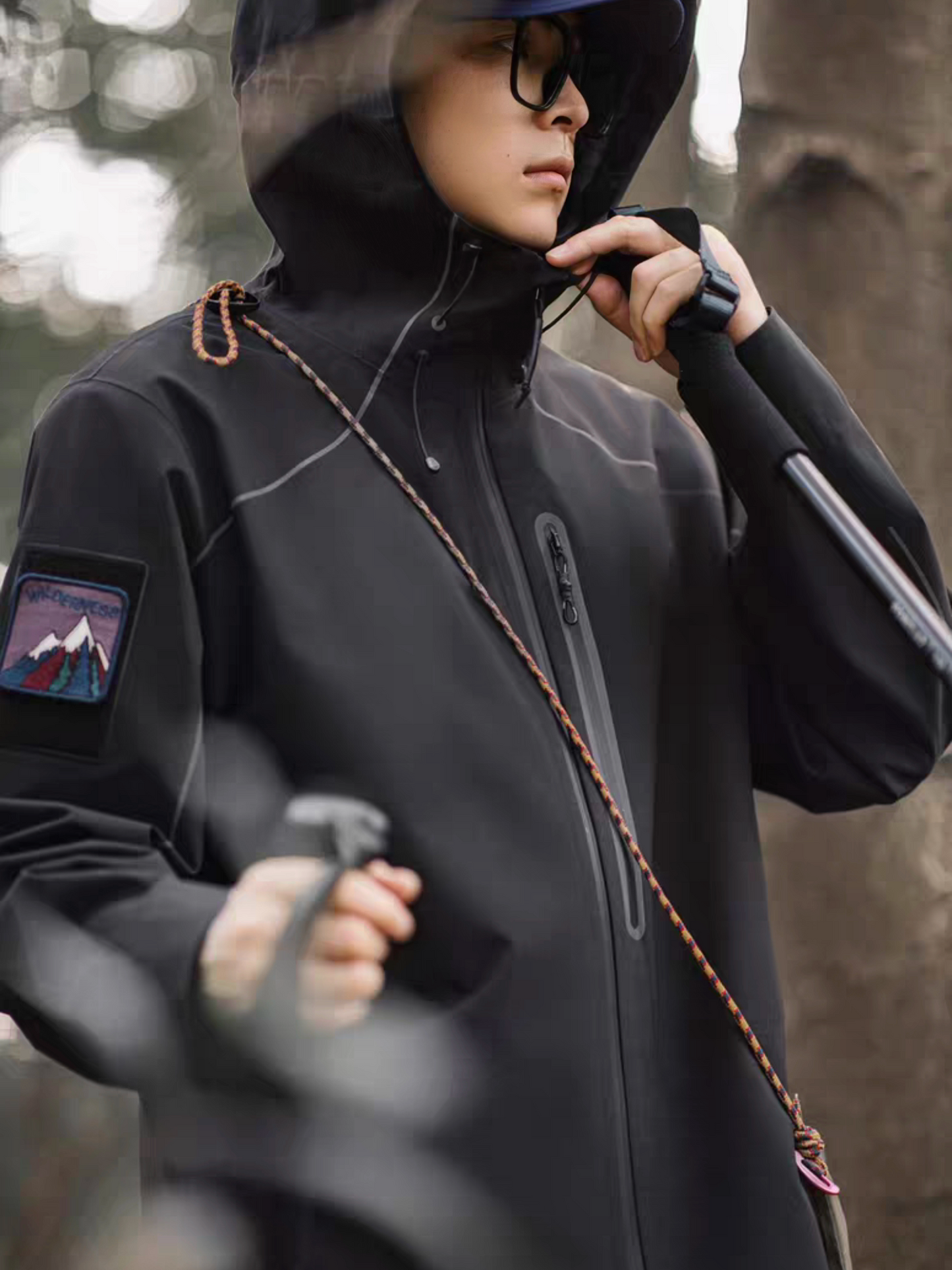 Close-up side view of model wearing STVLE lightsaber techwear jacket, showcasing mountain patch on sleeve, hiking rope accessory, and waterproof zipper in outdoor forest setting