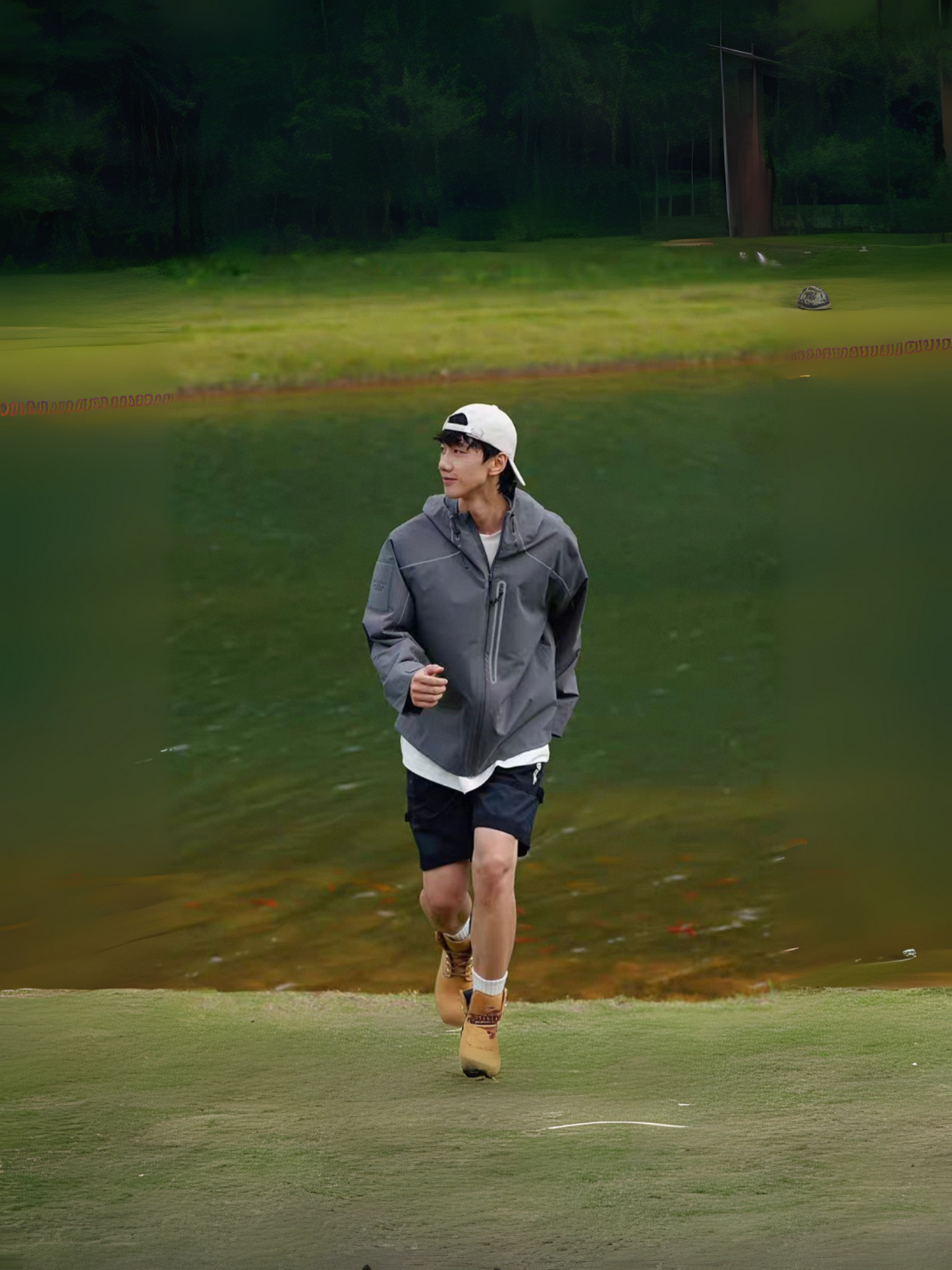 Model jogging outdoors on grass near a lake, wearing STVLE lightsaber techwear jacket with hiking boots and shorts, demonstrating outdoor functionality and breathability