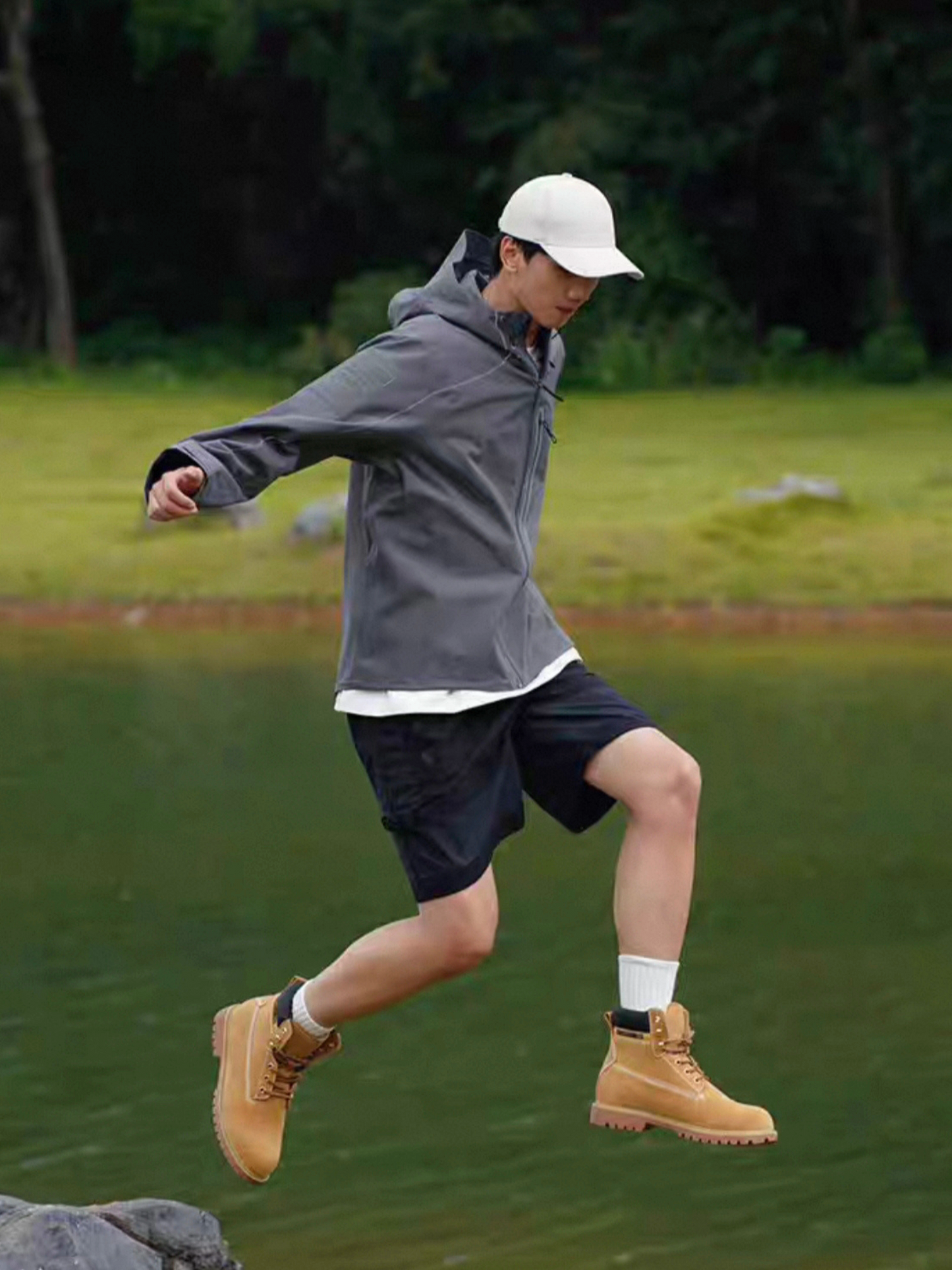 Dynamic outdoor shot of model mid-air jumping over rocks, wearing STVLE lightsaber techwear jacket with shorts and boots, demonstrating the jacket's mobility and functional performance