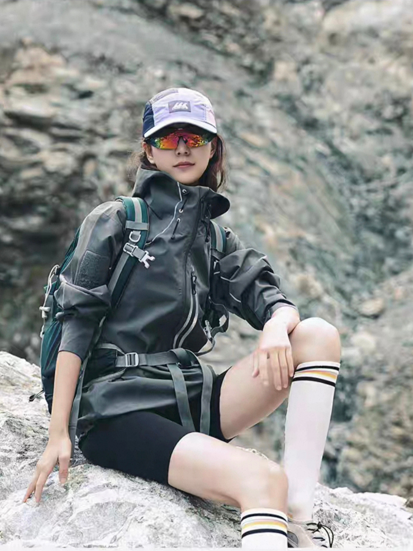 Female model sitting on a rock wearing STVLE lightsaber techwear jacket with a hiking backpack, sporty sunglasses and knee-high socks, showcasing a unisex mountain-inspired outdoor look