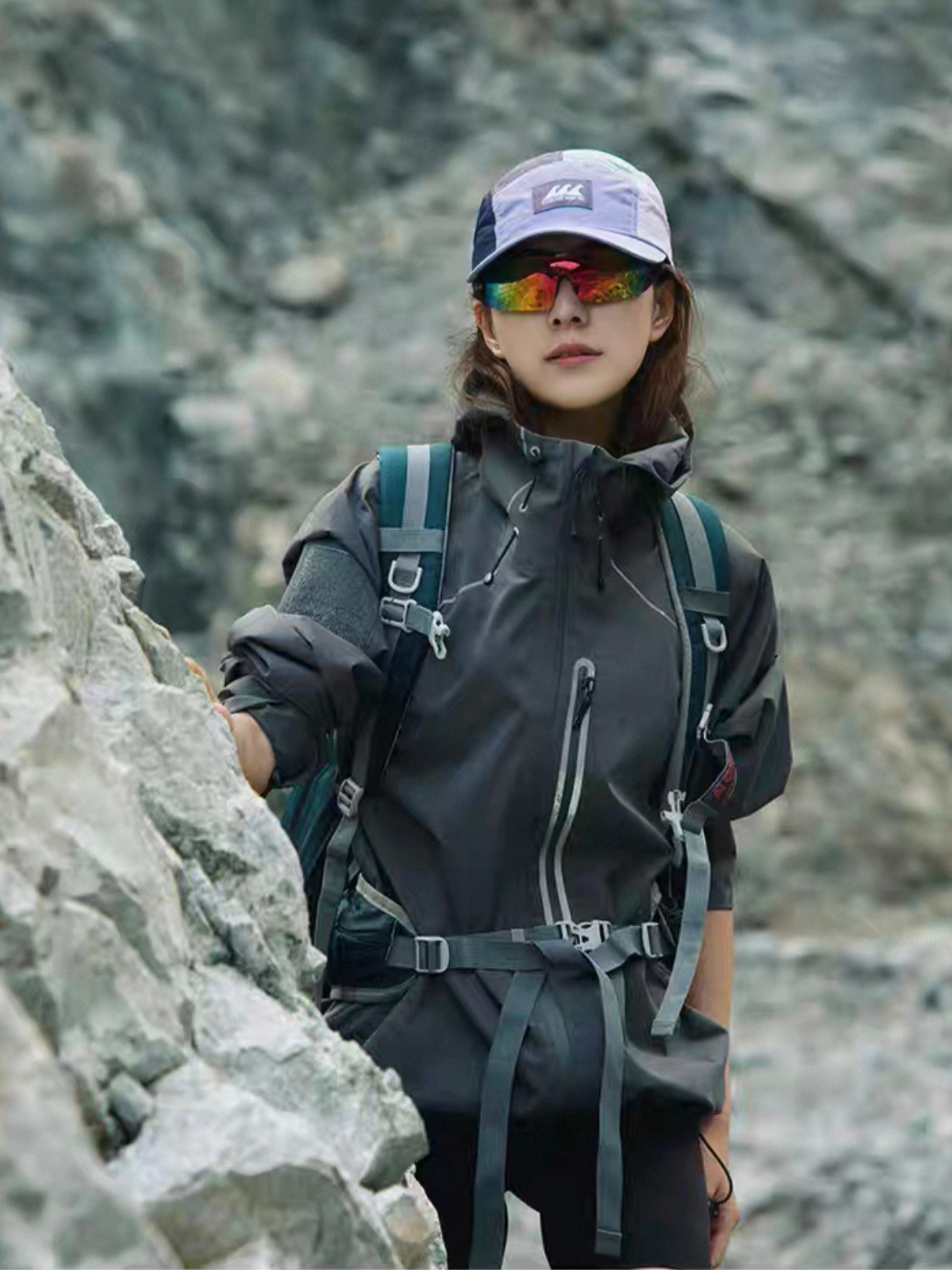 Female model standing in front of rocky mountain wall, wearing STVLE lightsaber techwear jacket and hiking gear, showcasing waterproof functionality and bold outdoor fashion