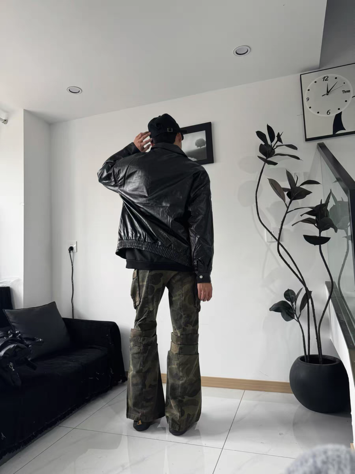 Male model showcasing back view of black PU varsity jacket with camo flared pants and cap. Indoor streetwear style with modern attitude