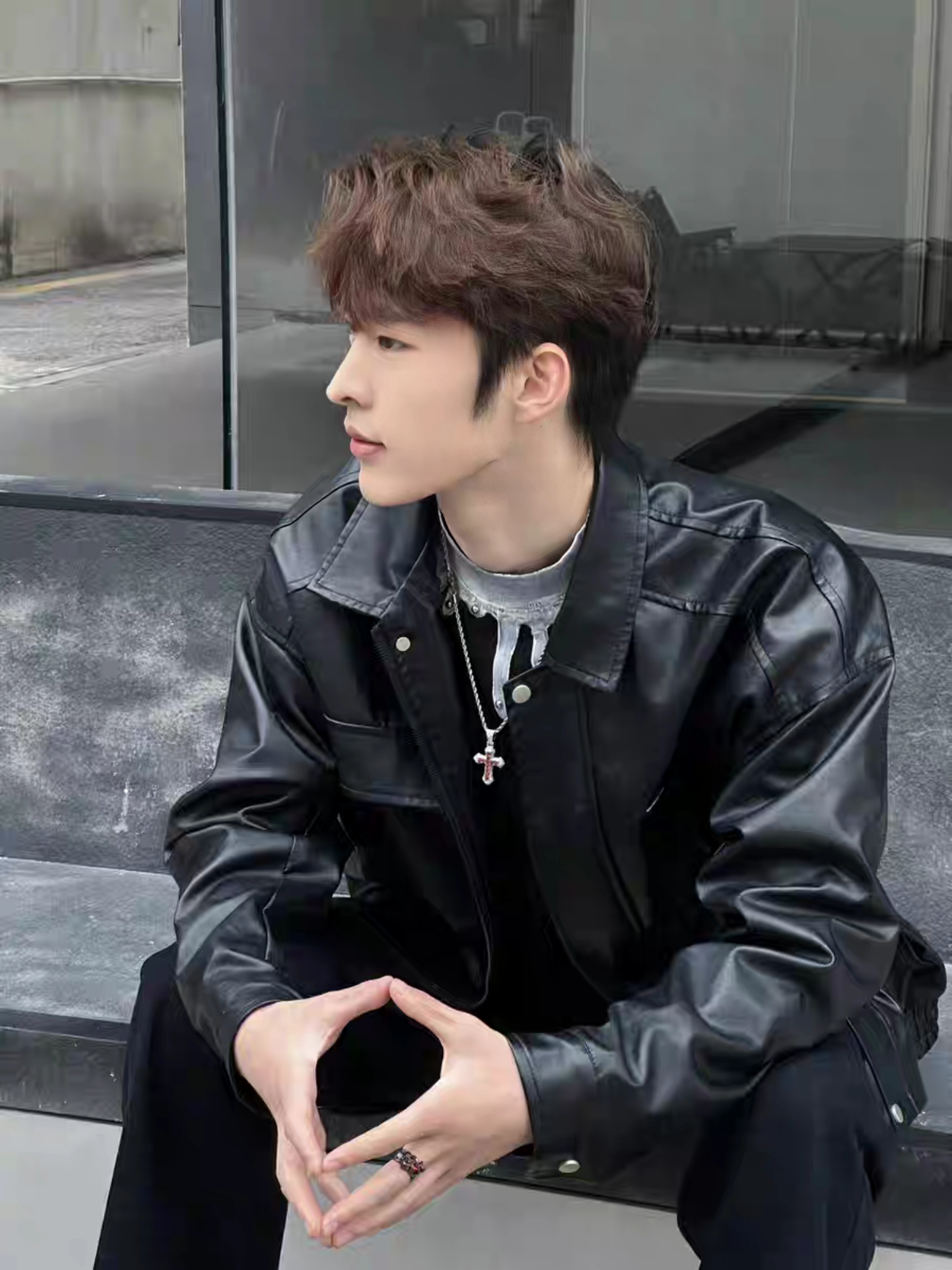 Male model in black faux leather jacket with silver cross necklace , sitting on a concrete bench outdoors showing side profile, K-style street fashion