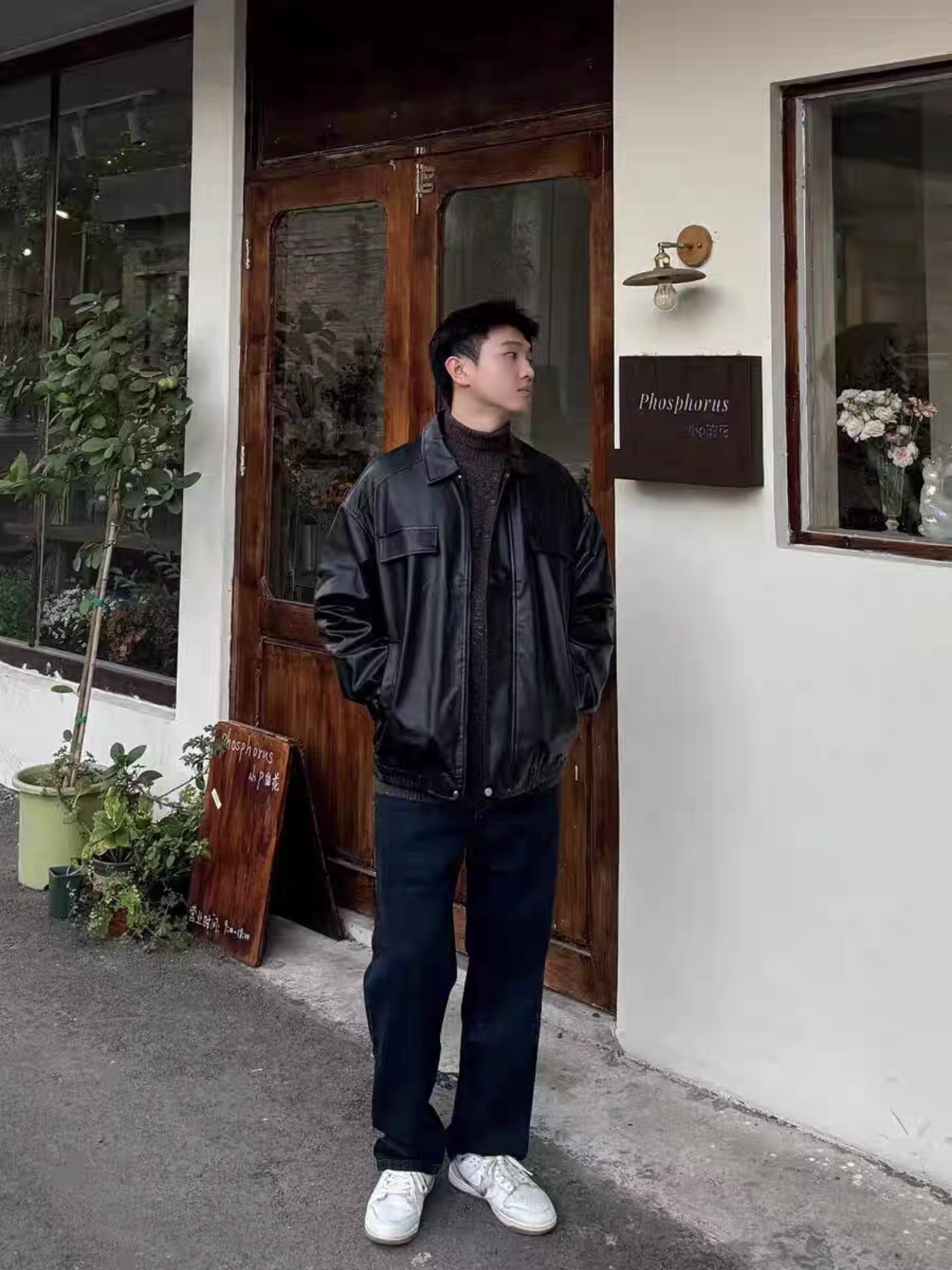 Male model wearing black PU varsity jacket standing outside a cafe, styled with dark jeans and white sneakers. Urban casual outfit inspiration.