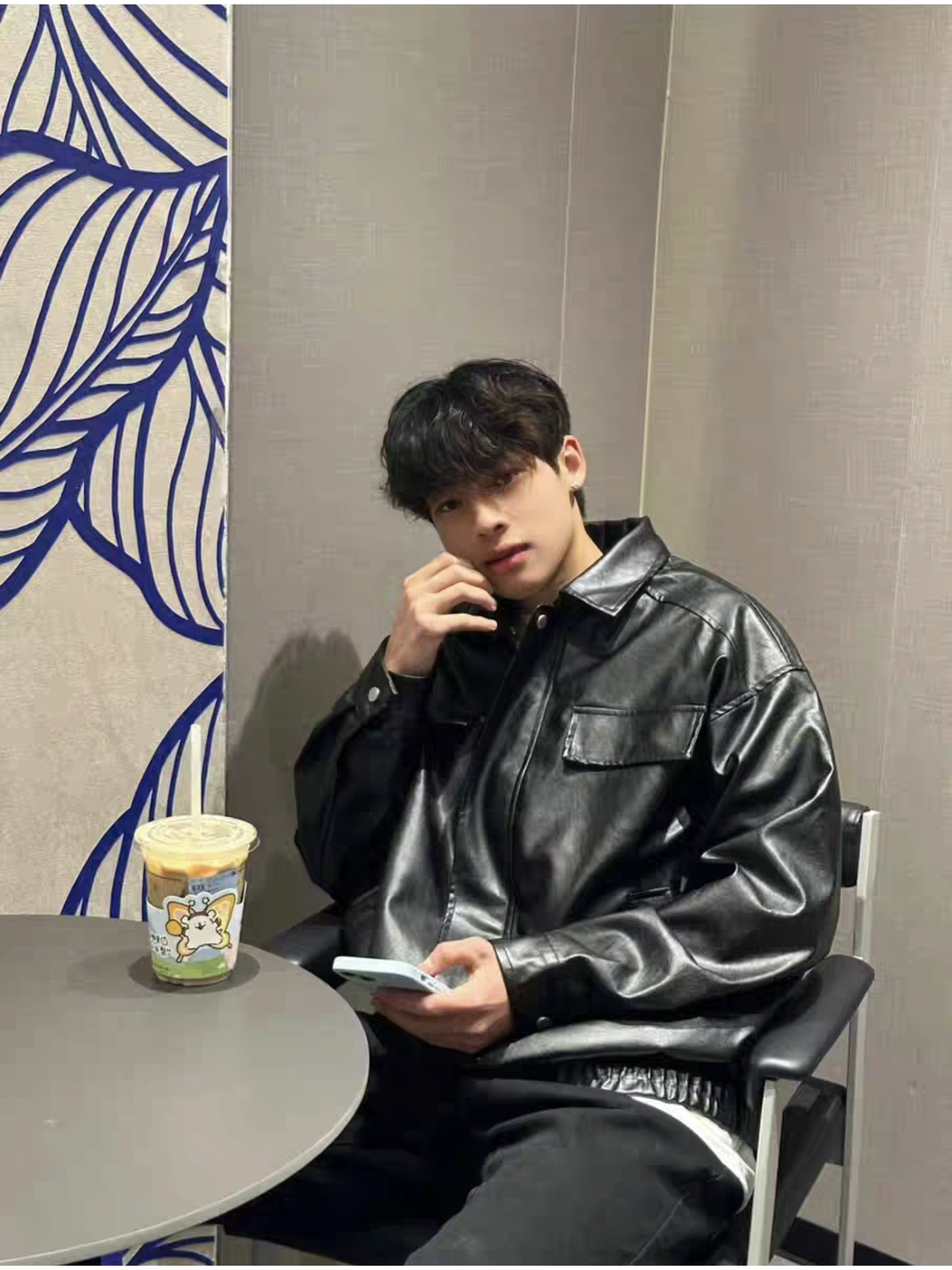 Asian male model sitting in cafe wearing STVLE black PU varsity jacket, styled casually with layered shirt and black pants, holding a phone with coffee on the table