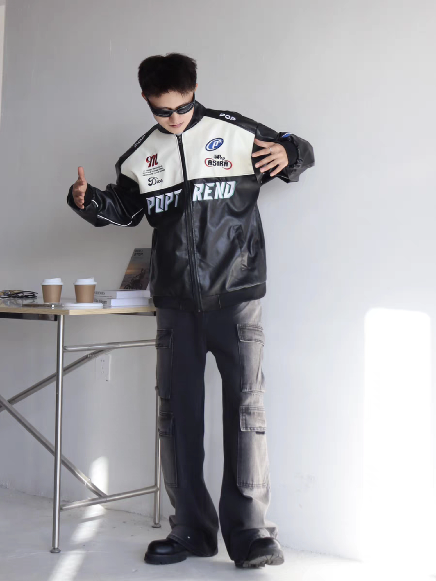 Male model in black racing-style PU leather jacket with embroidered acing patches, striking a bold pose indoors with sunglasses and cargo pants