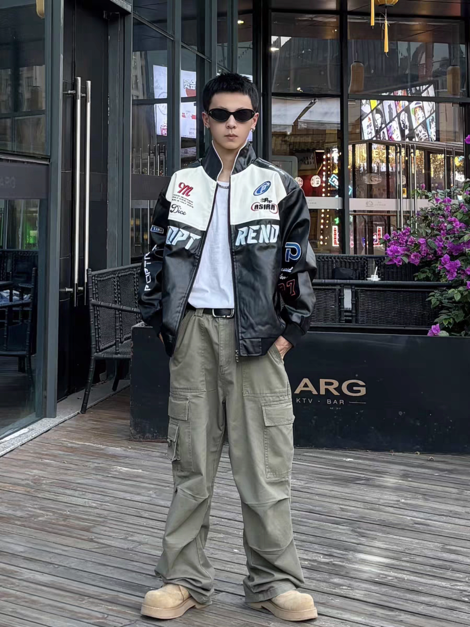 Male model standing outside a stylish bar wearing black racing PU leather jacket with sunglasses and cargo pants, showing nightlife streetwear lookj