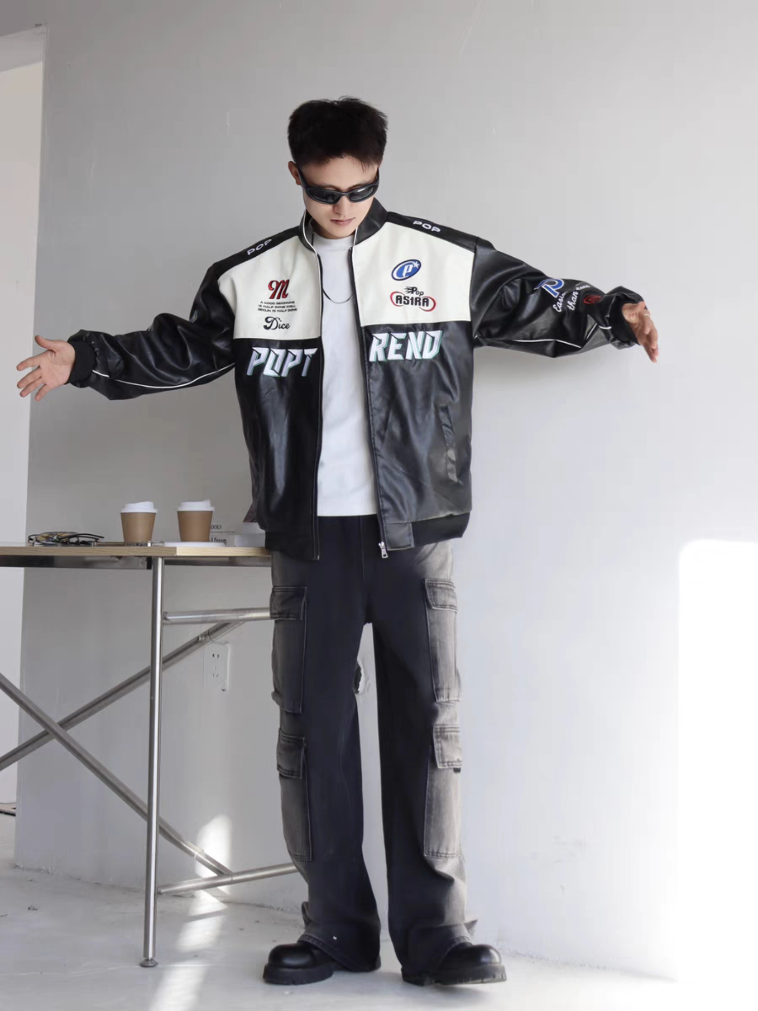 Male model wearing black racing PU jacket with arms open, showcasing relaxed fit and embroidered front details urban indoor setting