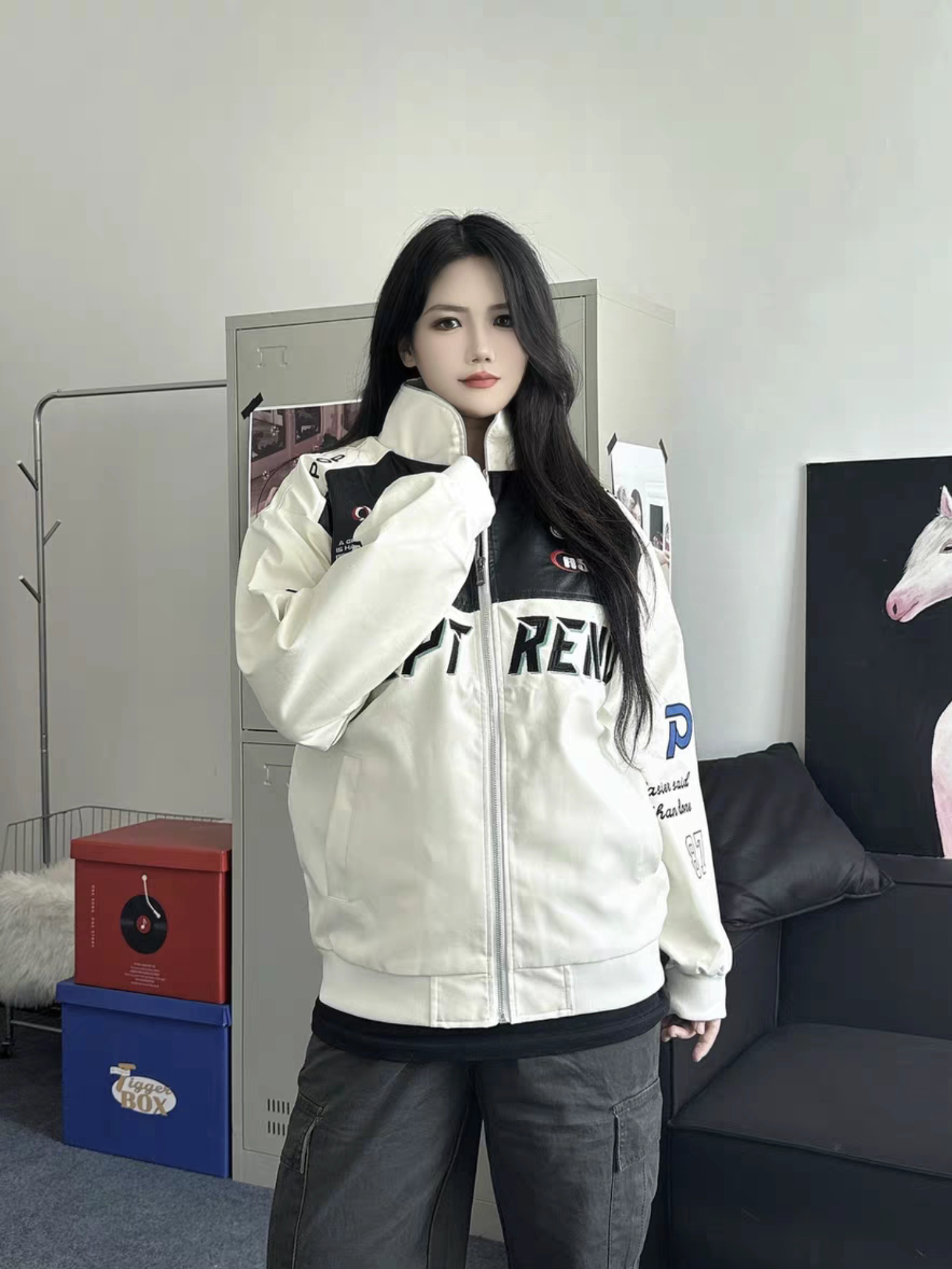 Female model wearing a white racing style moto jacket with embroidered logos, standing indoors in a minimalistic room with lockers and modern decor
