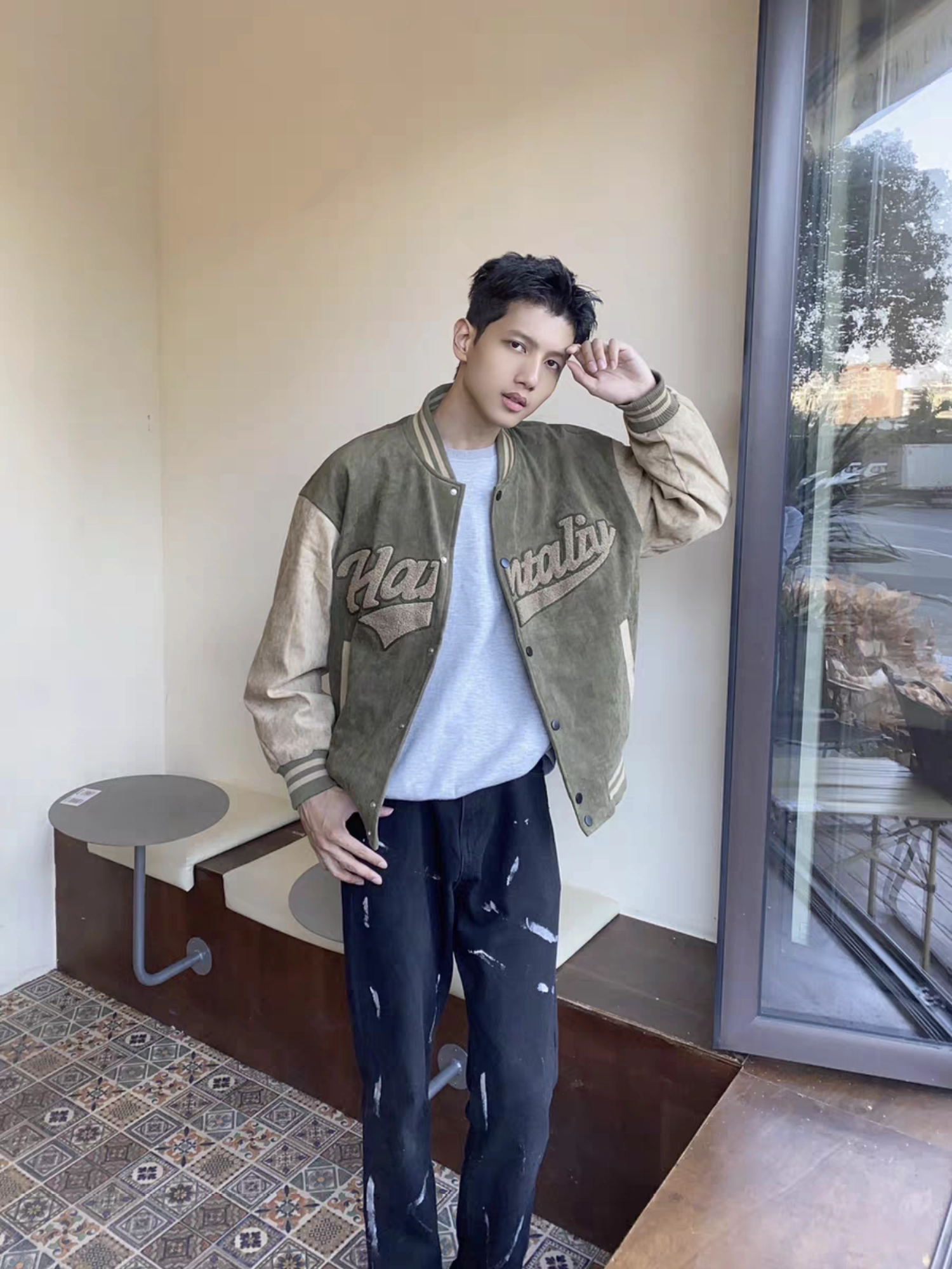 Male model posing in green and brown Hantaliv jacket next to a window, paired with distressed black jeans and gray tee in a lifestyle setting