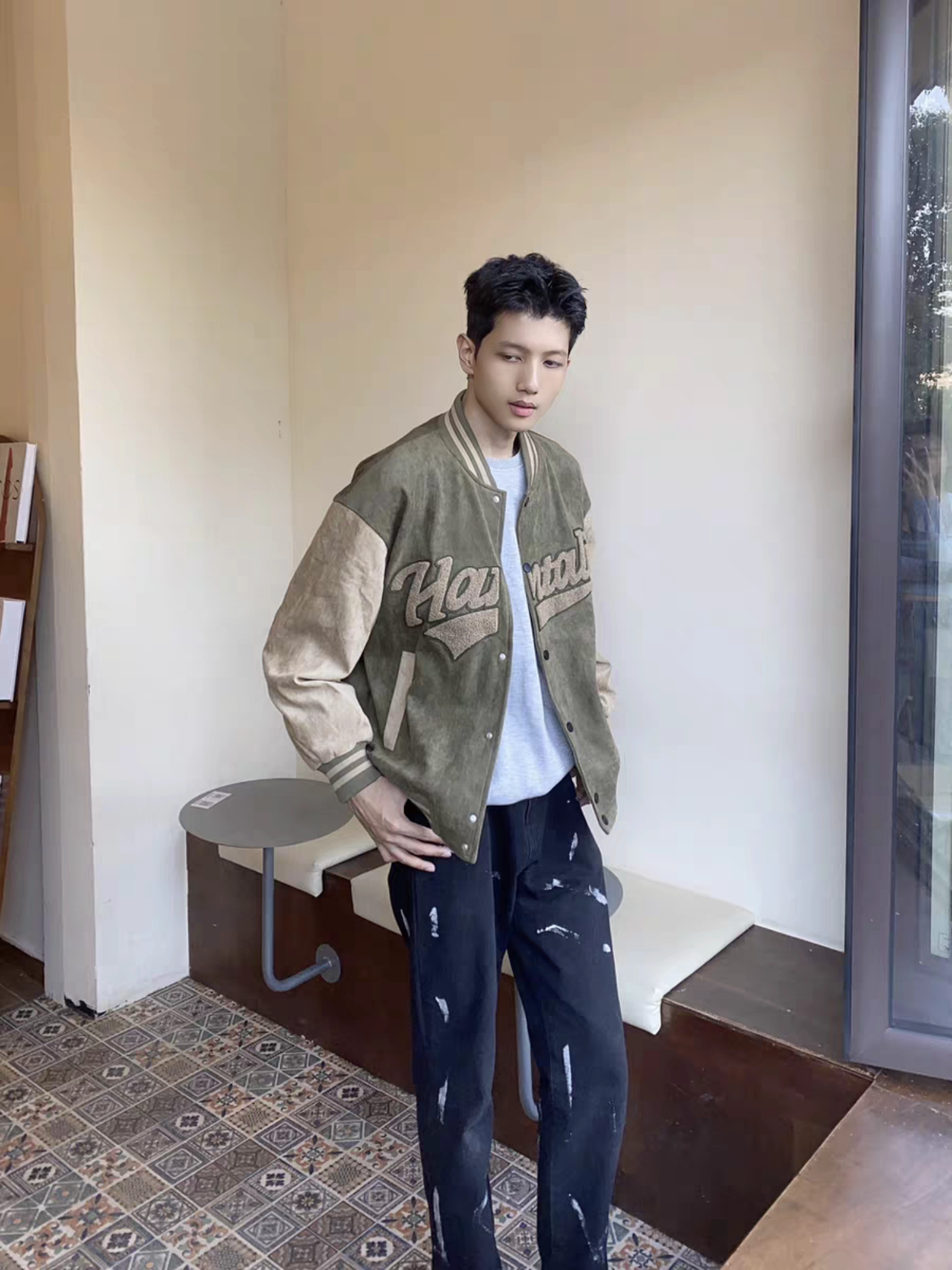 Male model wearing green and brown Hantaliv varsity jacket standing indoors near a window, styled with distressed black jeans and grey T-shirt