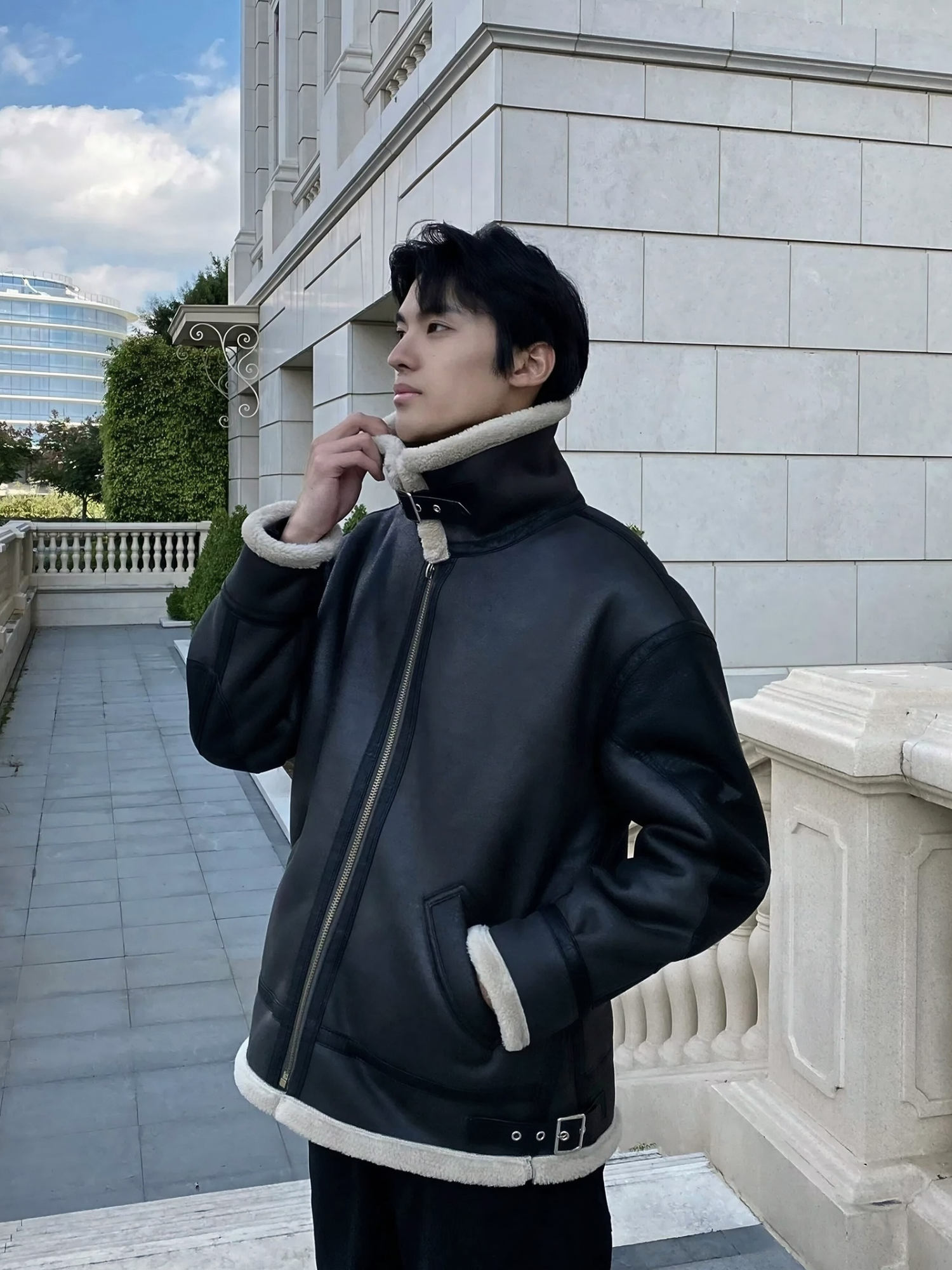 Man wearing black faux shearling aviator jacket with buckle strap collar, standing in front of European-style building