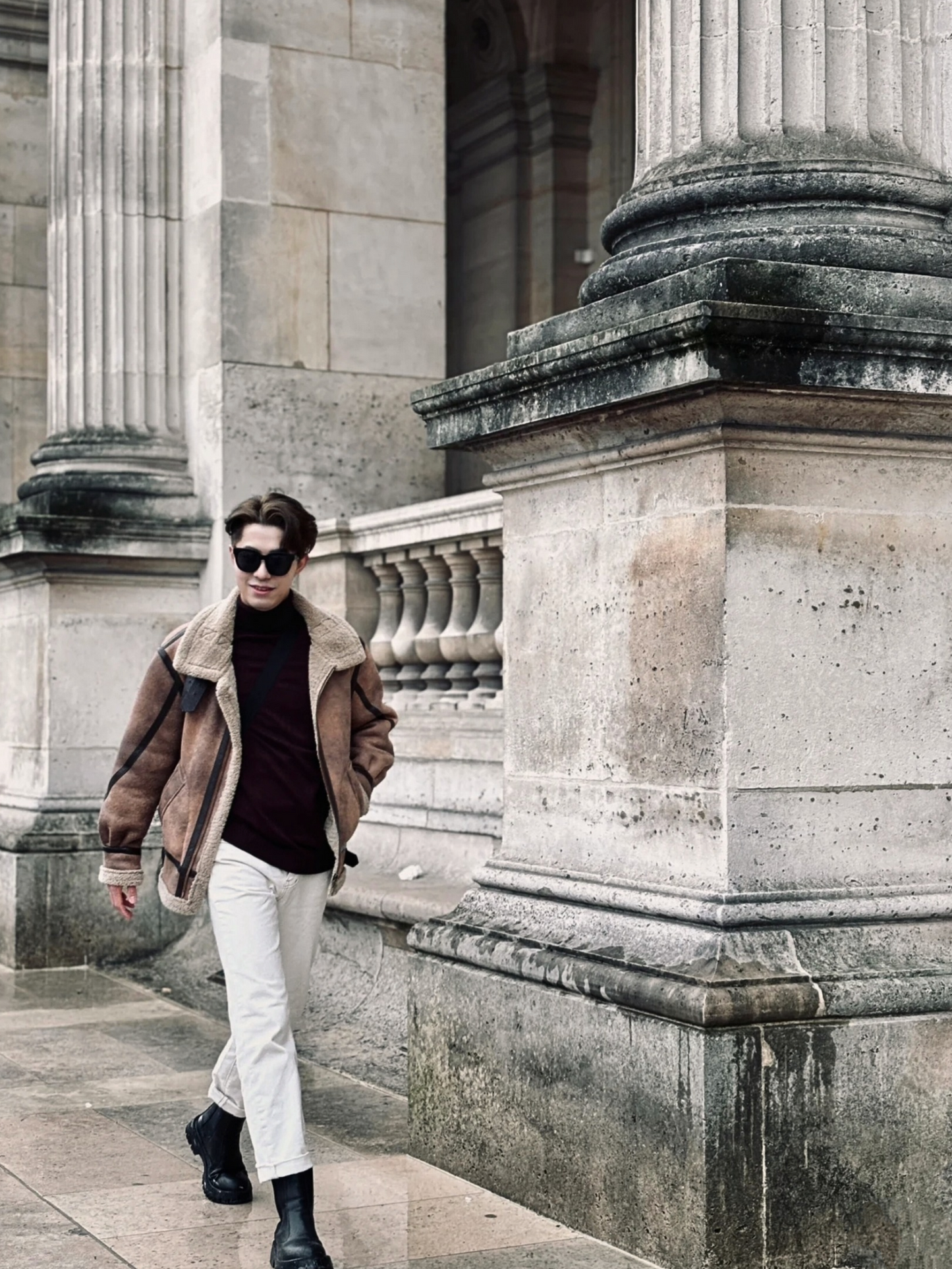 Man wearing brown faux shearling aviator jacket, walking near classical European architecture in winter outfit