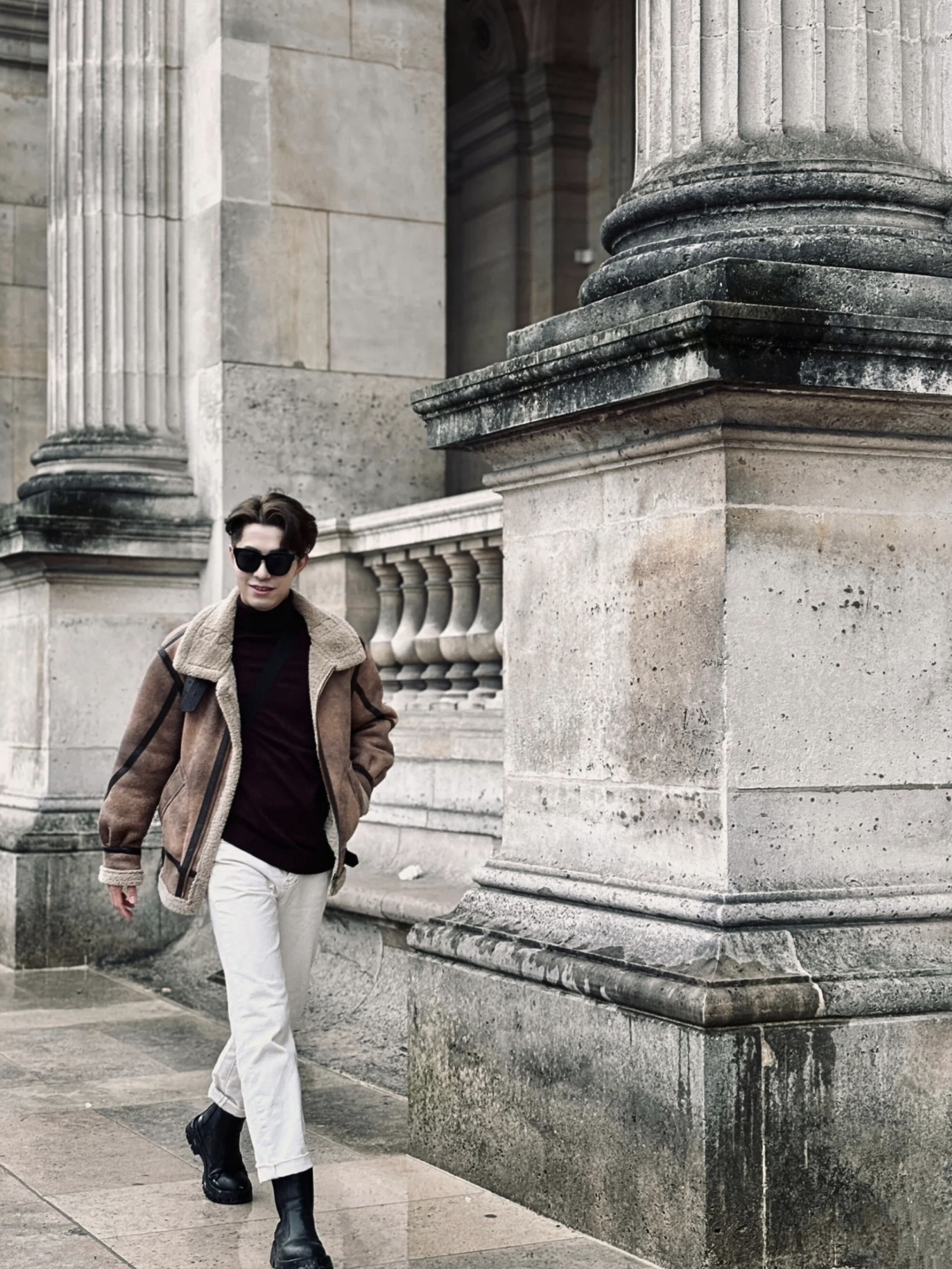 Man wearing brown faux shearling aviator jacket, walking near classical European architecture in winter outfit