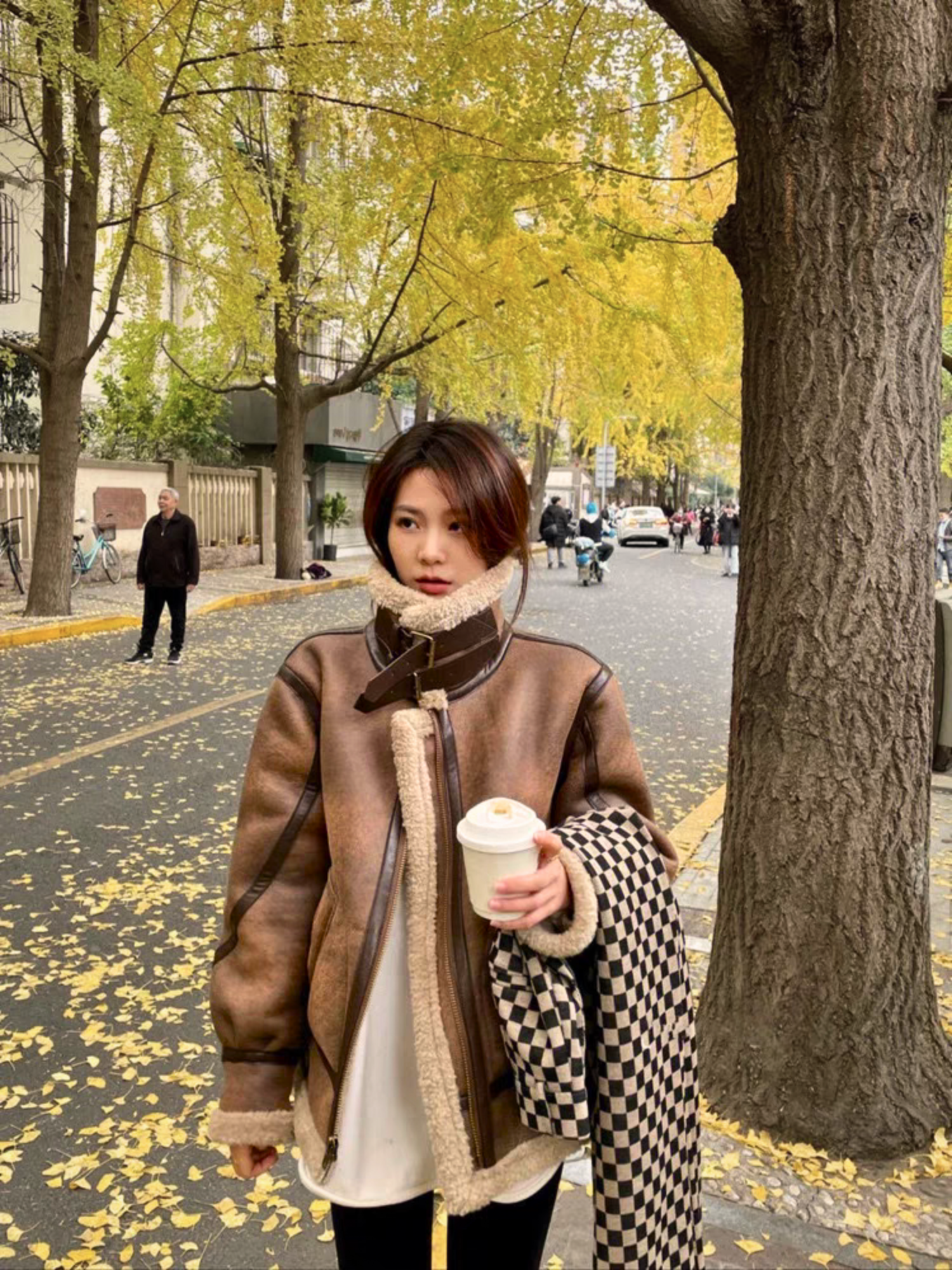 Woman wearing brown faux shearling aviator jacket holding coffee cup on autumn street with ginkgo leaves