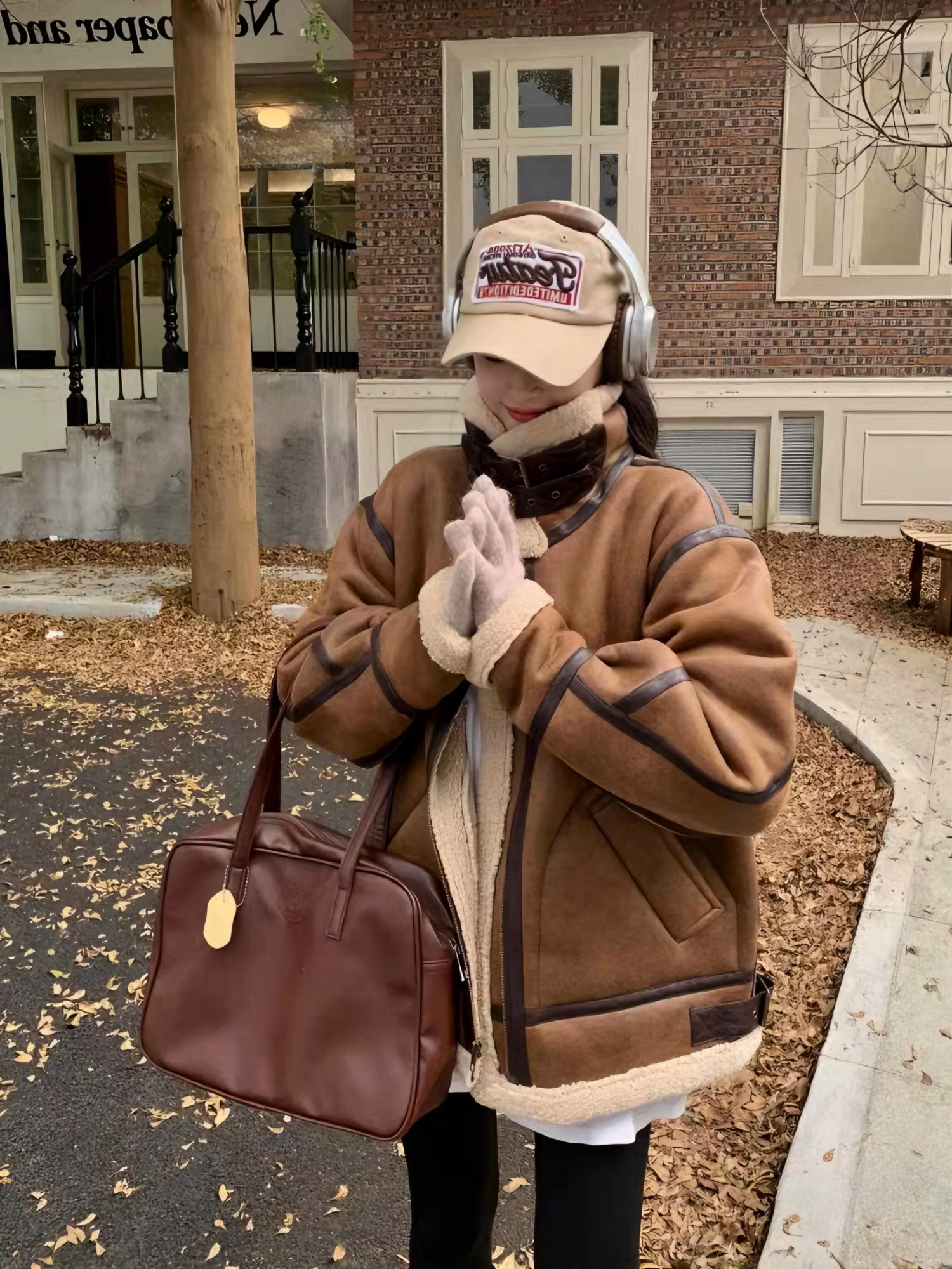 Woman in brown faux shearling aviator jacket with headphones and cap, holding hands together in autumn street setting