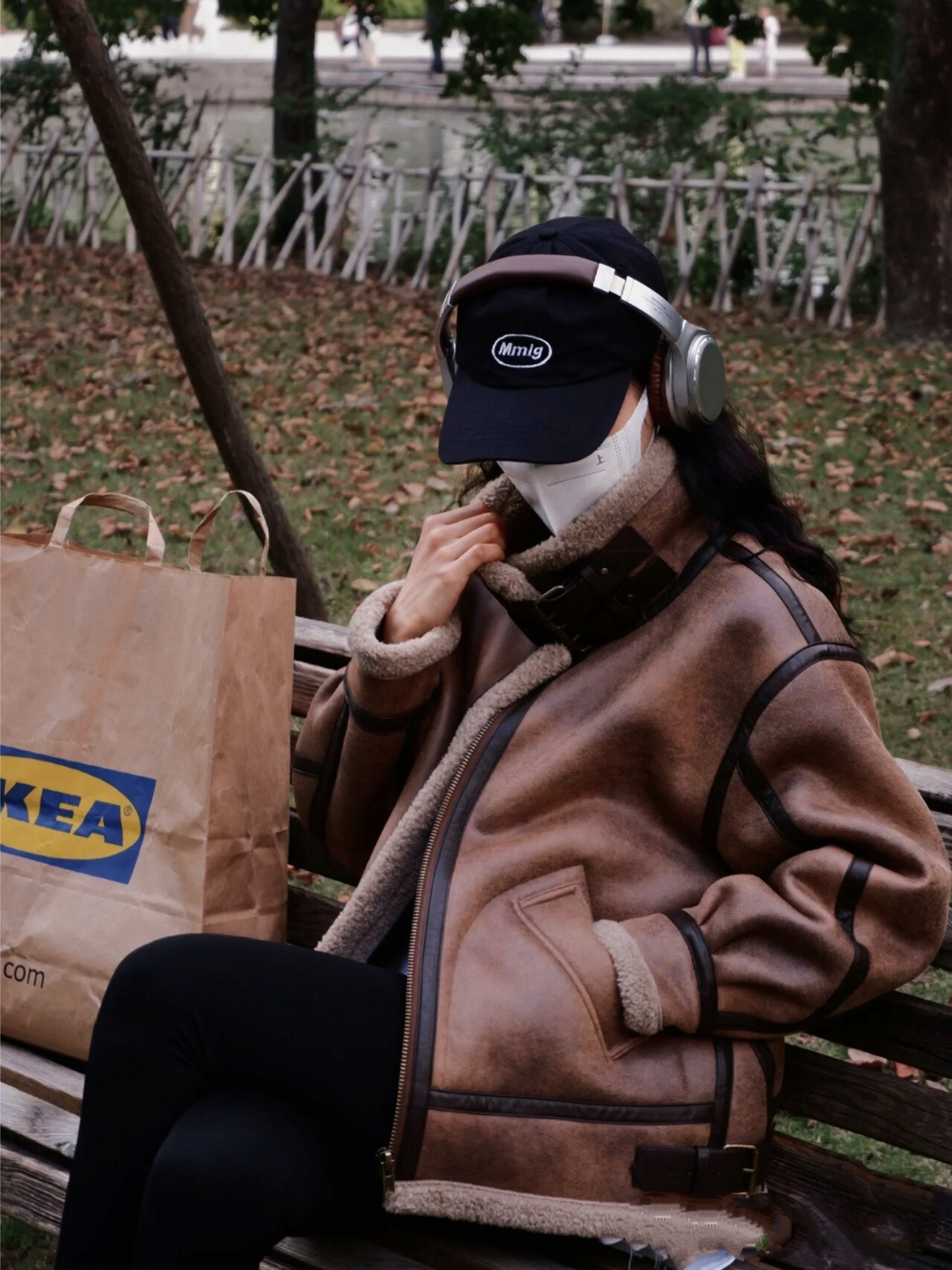 Woman in brown faux shearling aviator jacket with high collar, wearing cap and headphones, sitting on park bench with shopping bag