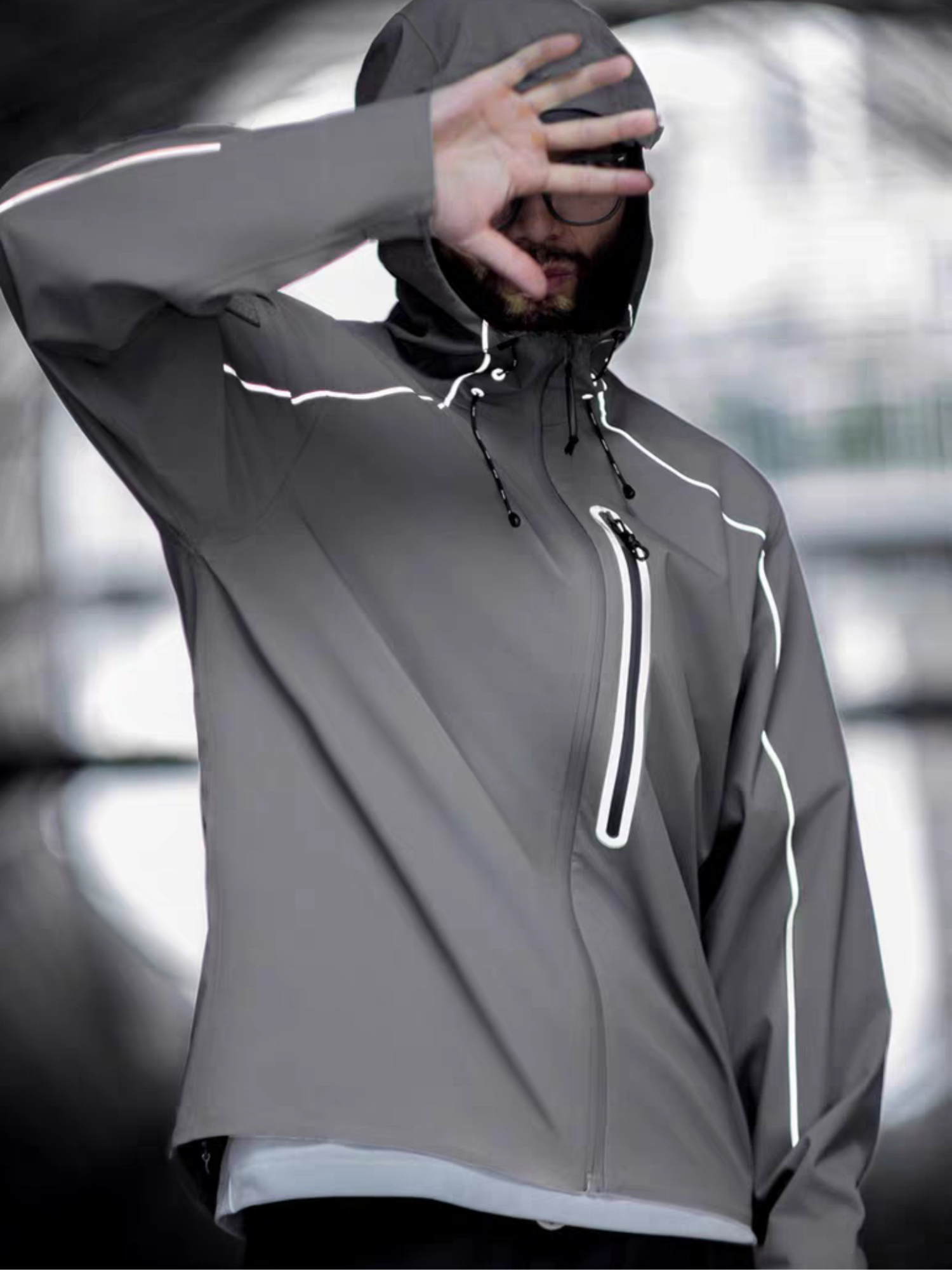 Model wearing STVLE lightsaber reflective techwear jacket, hood on, hand covering face - -shot under urban overpass with tactical posture and minimalist  design details