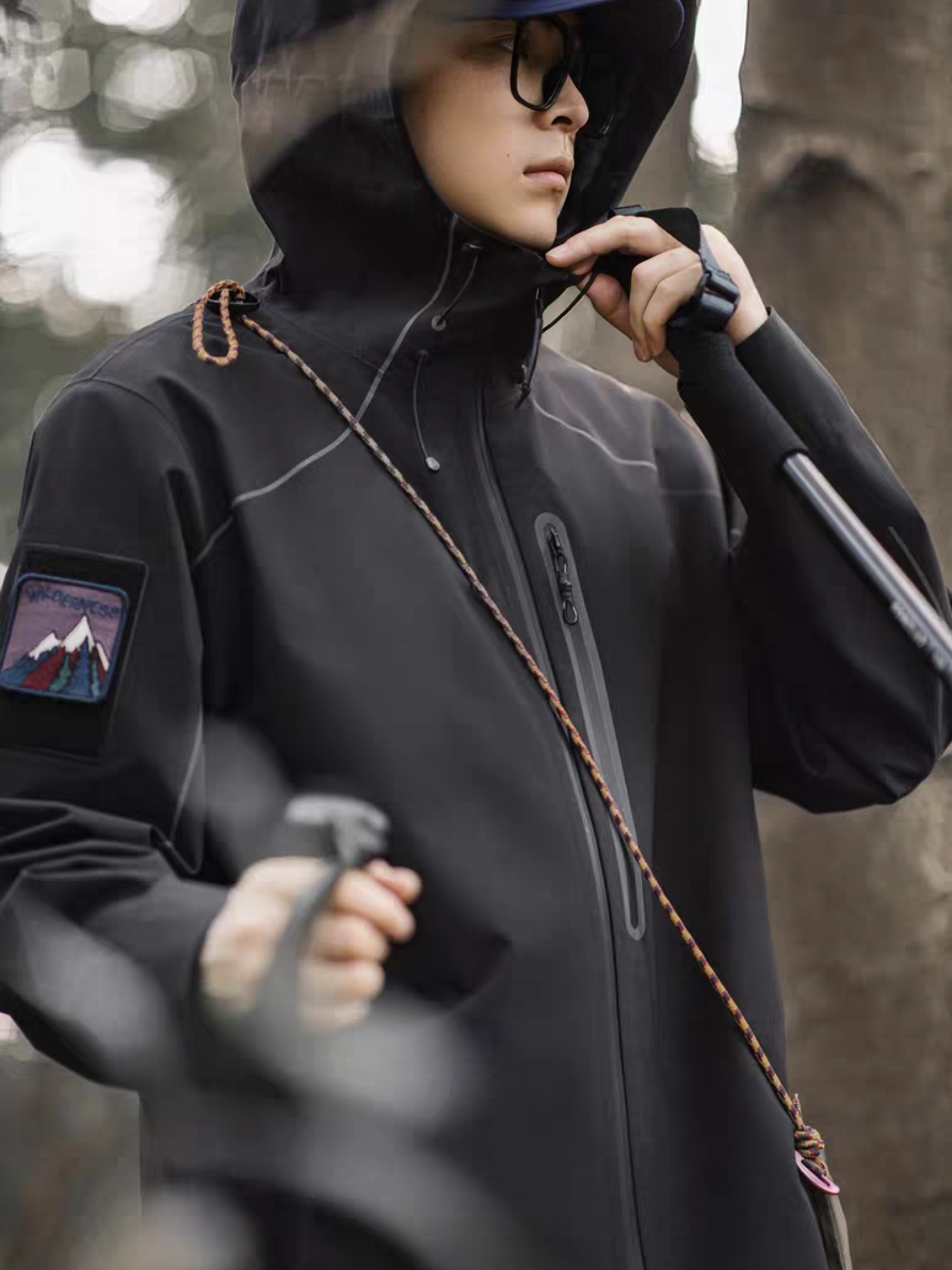 Close-up side view of model wearing STVLE lightsaber techwear jacket, showcasing mountain patch on sleeve, hiking rope accessory, and waterproof zipper in outdoor forest setting
