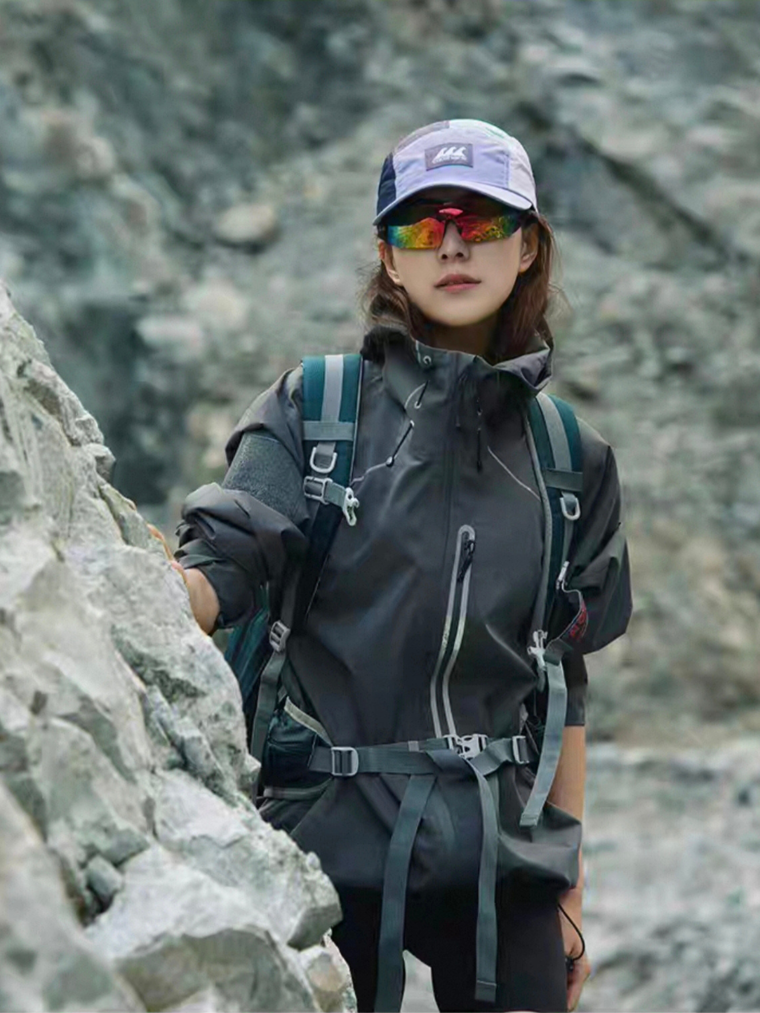 Female model standing in front of rocky mountain wall, wearing STVLE lightsaber techwear jacket and hiking gear, showcasing waterproof functionality and bold outdoor fashion